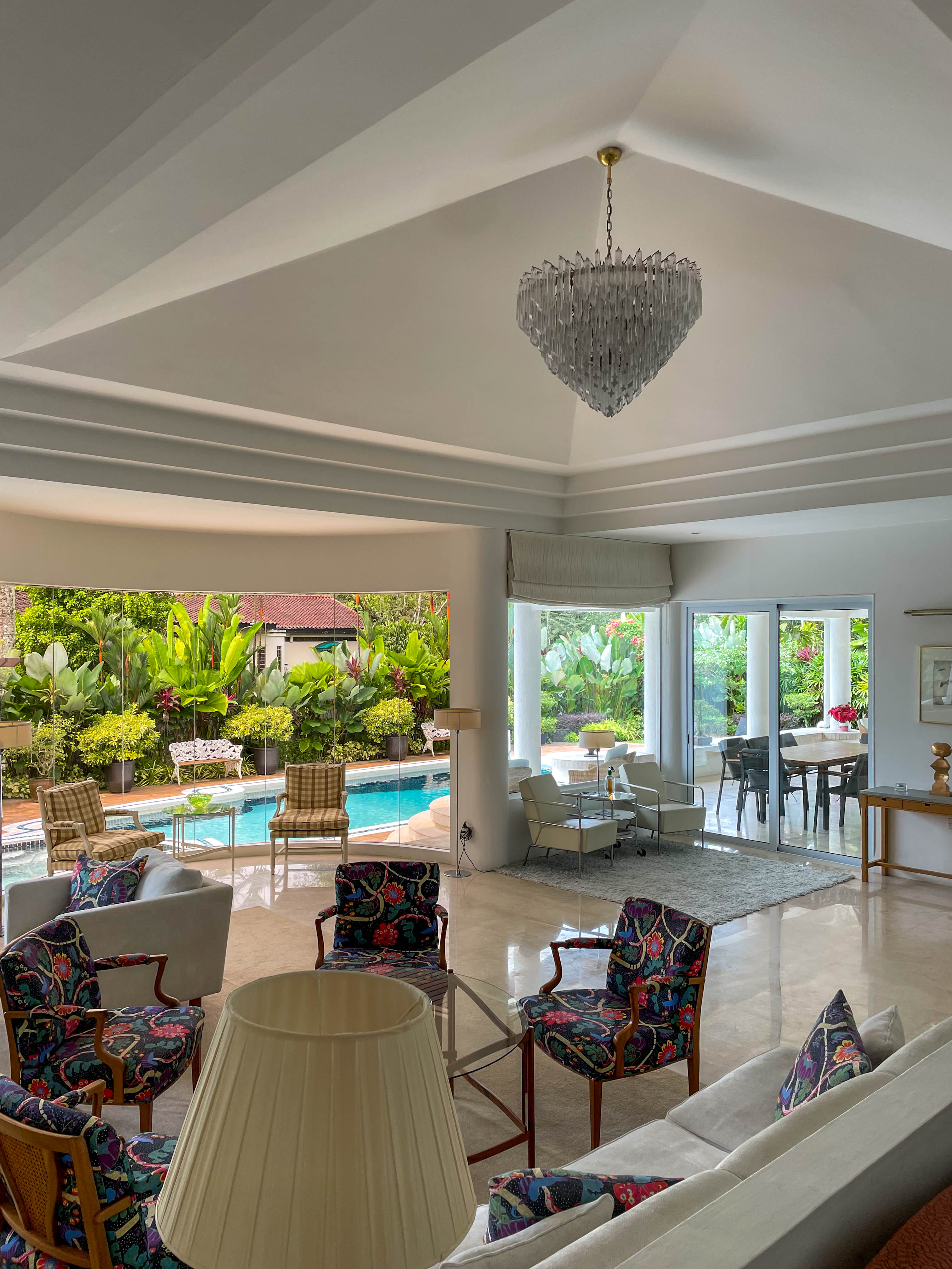 Ett ljust vardagsrum med högt i tak och kristallkrona. Nätta fåtöljer i färgglada mönster i gruppen. Utsikt mot en uteplats med pool