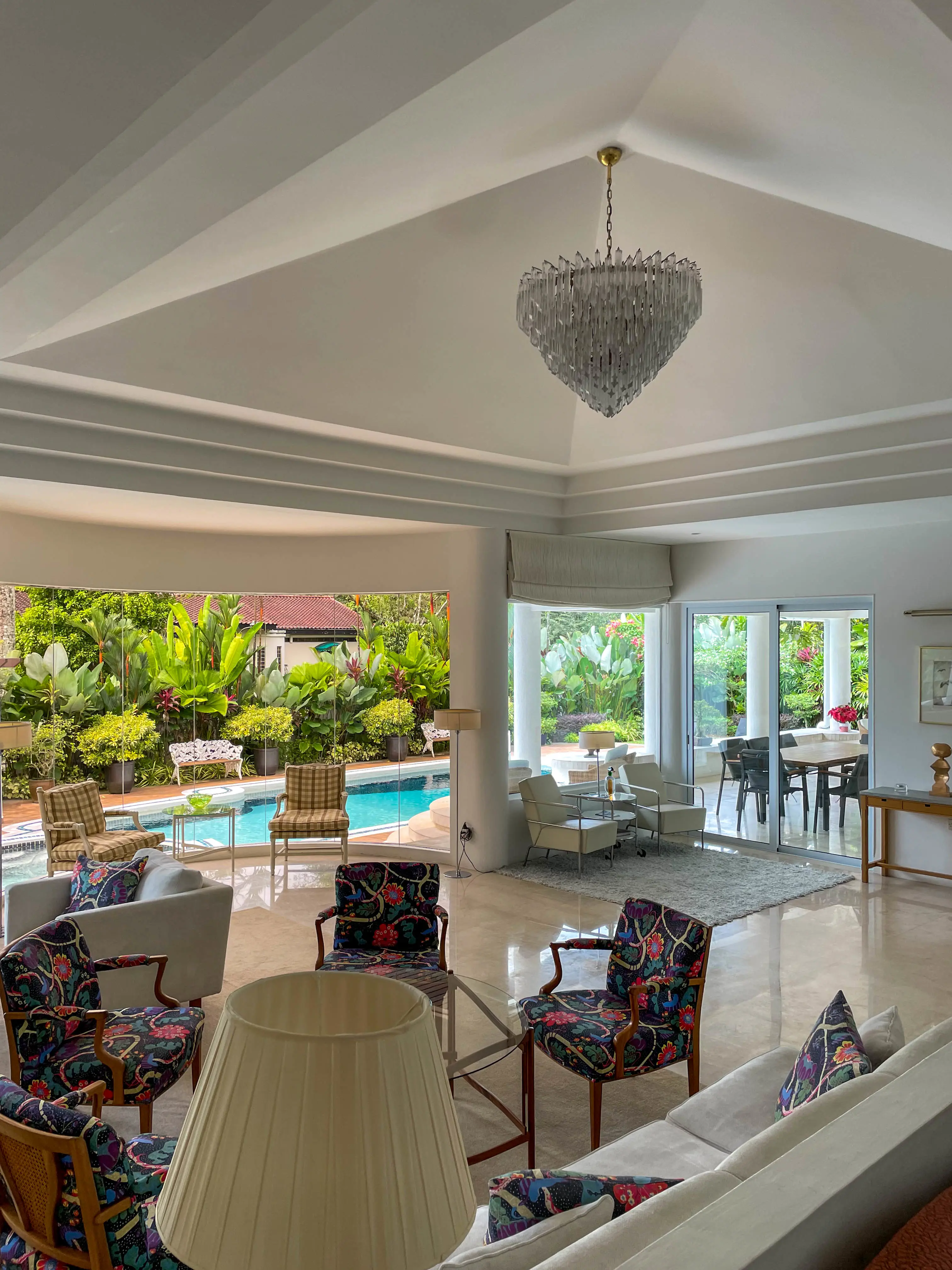 Ett ljust vardagsrum med högt i tak och kristallkrona. Nätta fåtöljer i färgglada mönster i gruppen. Utsikt mot en uteplats med pool