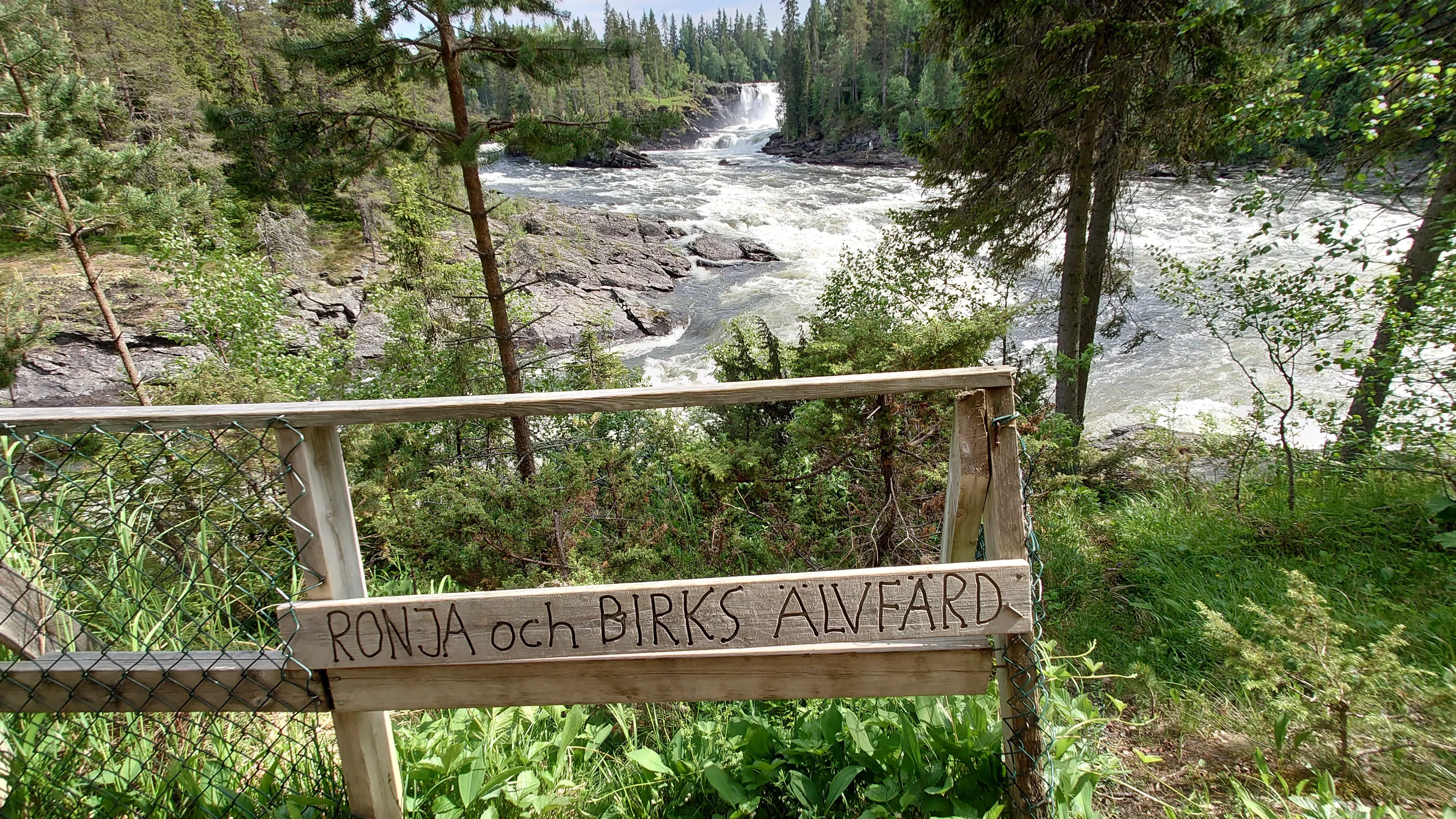 En forsande älv i granskog. På ett staket sitter en trä-skylt där det står "Ronja och Birks älvfärd".