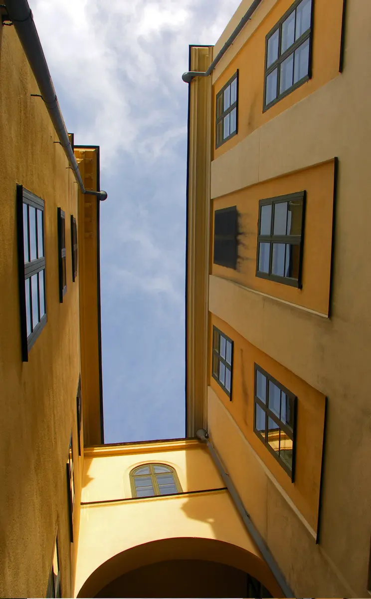 Gränd mellan två gulputsade byggnader fotograferade nedifrån med blå himmel som bakgrund. Husen förbinds av en gång över Gymnasiegränd..