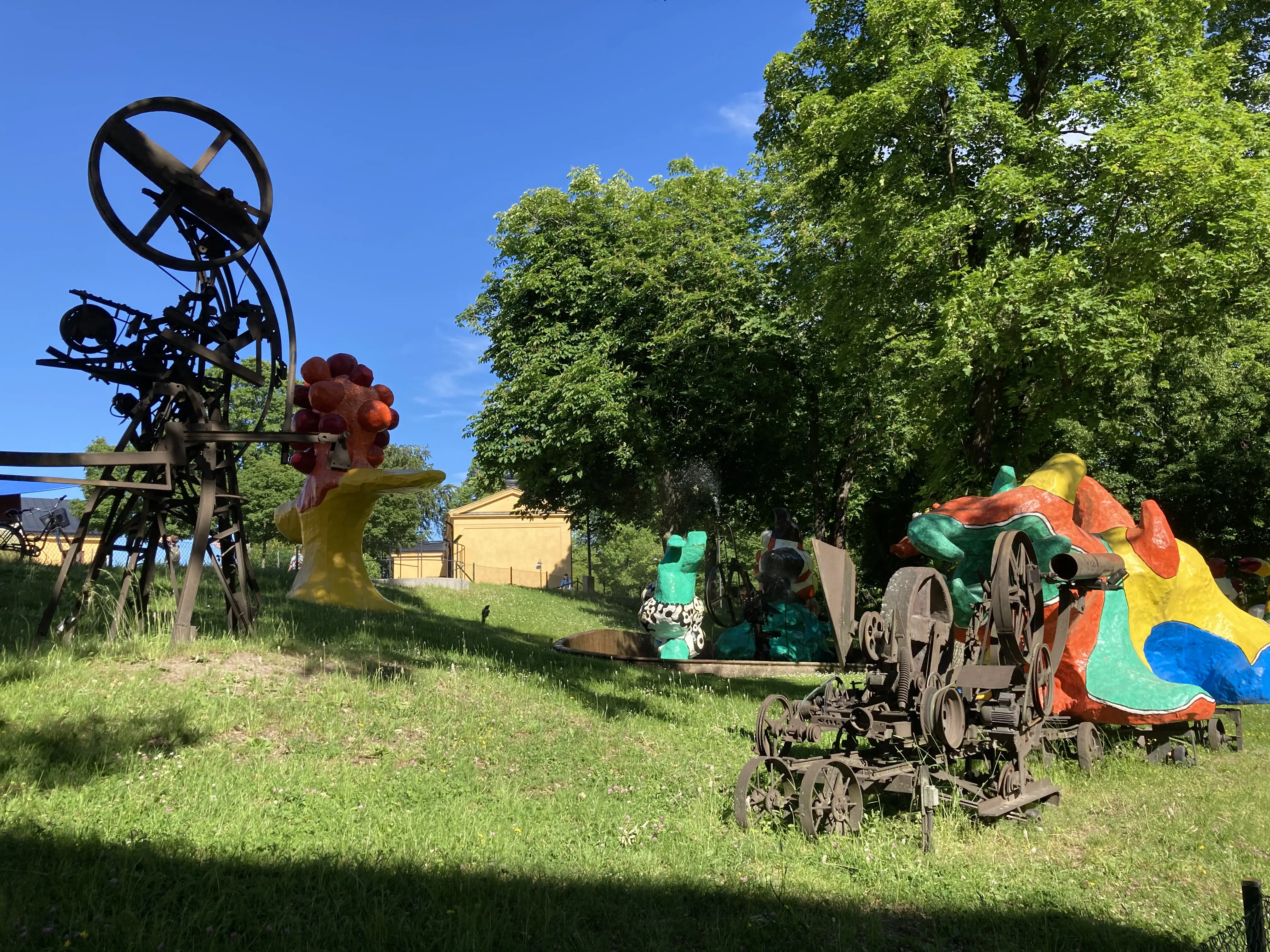 Skeppsholmen. Skulpturer på en gräsyta. Några är byggda av rostiga mekaniska delar, några är färg-glada med mjuka fomer.