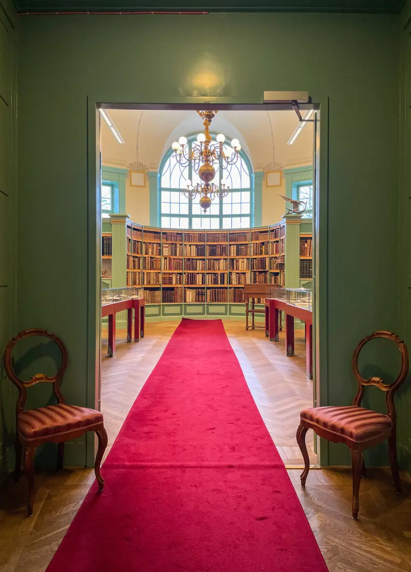 Dörröppning in till bibliotekssal med bokhyllor och en stor ljuskrona. En lång röd matta på trägolvet leder in i salen.
