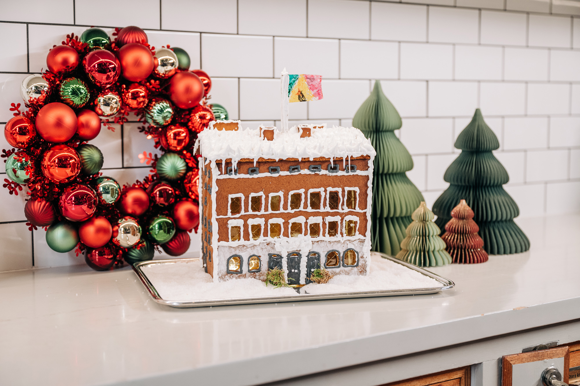 Ett dekorerat pepparkakshus står på en silvrig bricka på en köksbänk. Bakom den står en julkrans i rött och gröna pappersgranar.