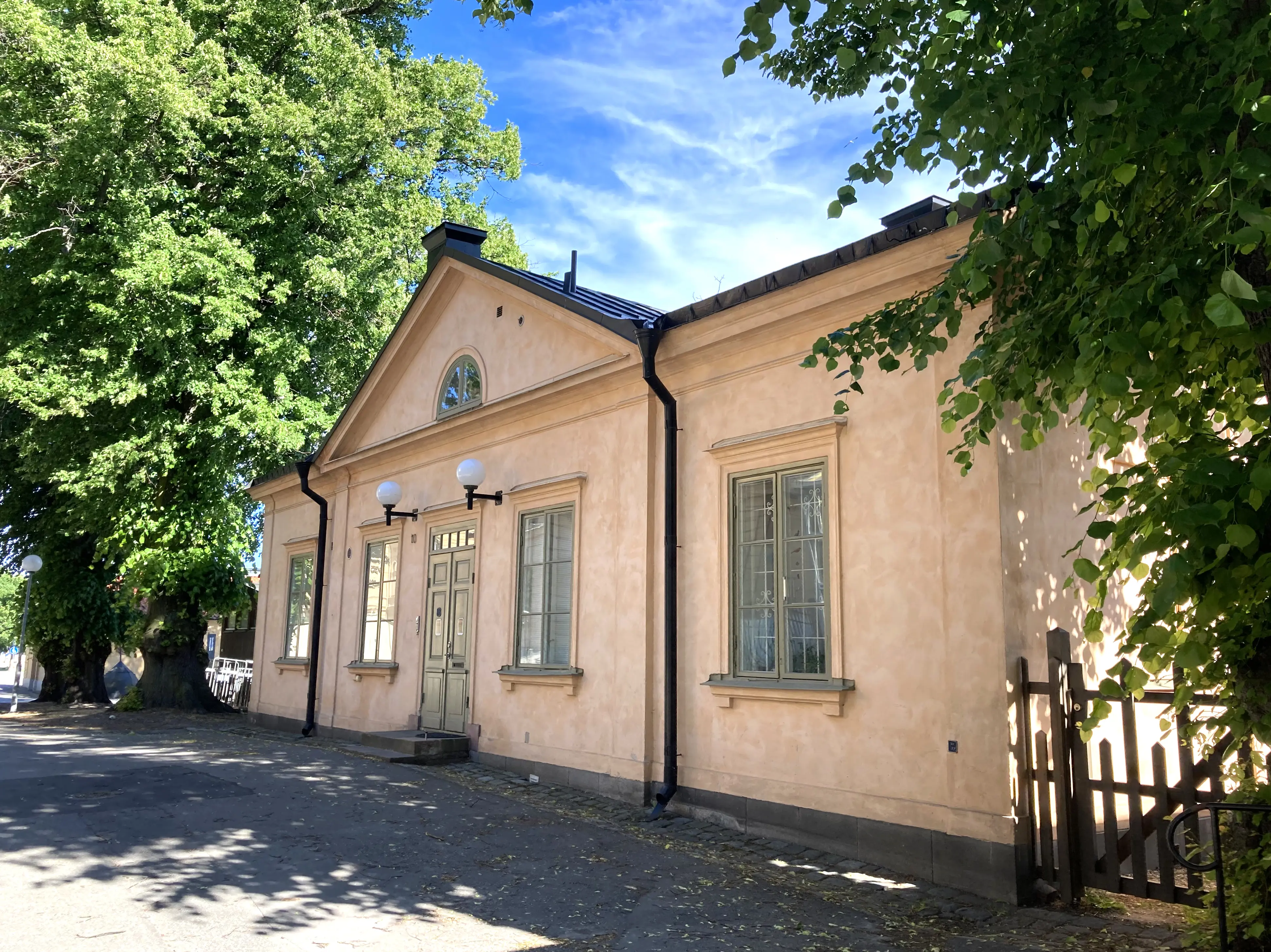 Kasernbefälhavarbostället. Klassicistisk liten byggnad i ljusrosa puts. En trägrind leder in till en trädgård. Sommar och grönska.