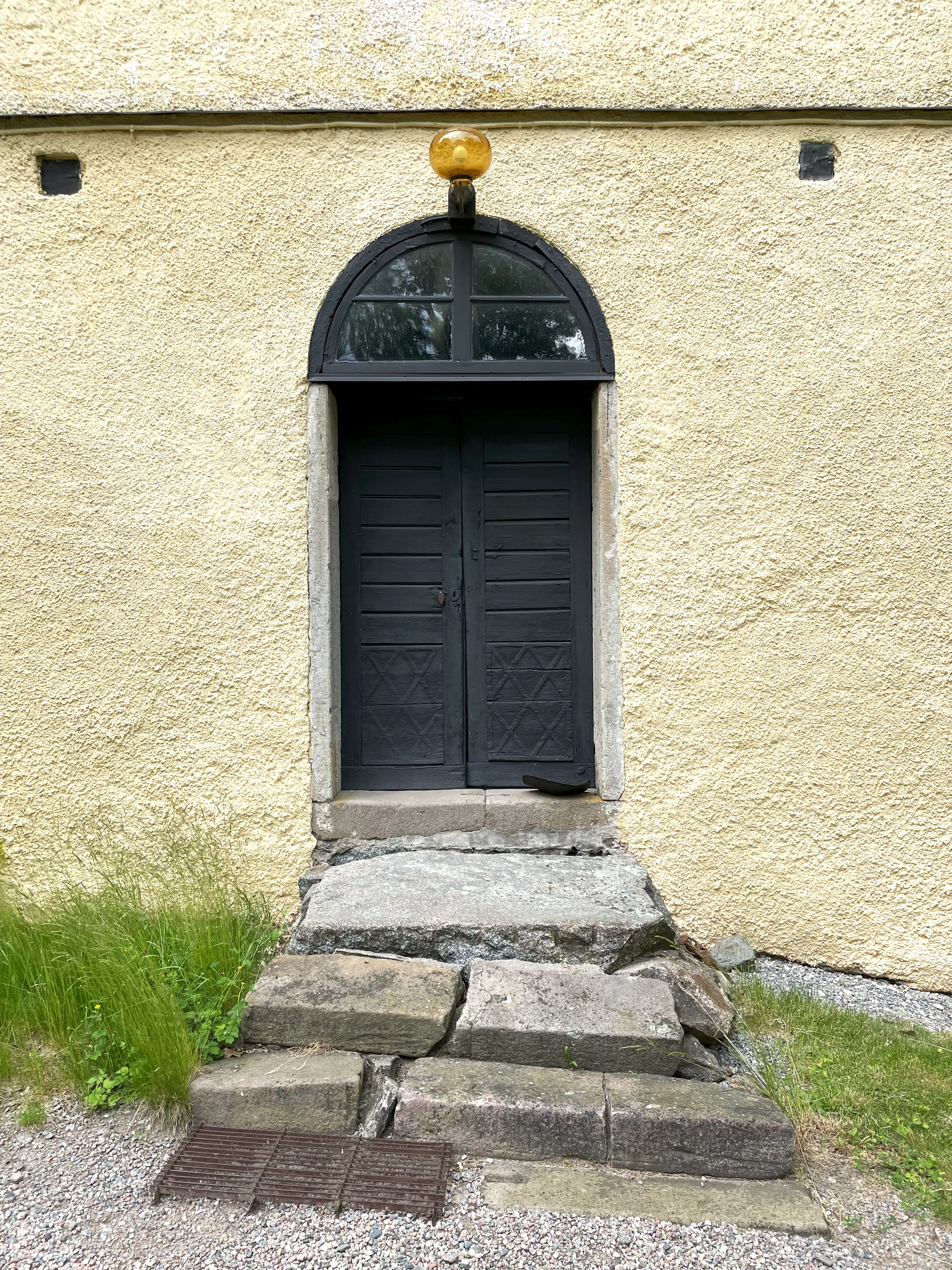 Landsberga. Sidodörr på huvudbyggnaden. En grov stentrappa leder upp till smala, svarta pardörrar. Ovanför sitter ett halvmåneformat fönster.