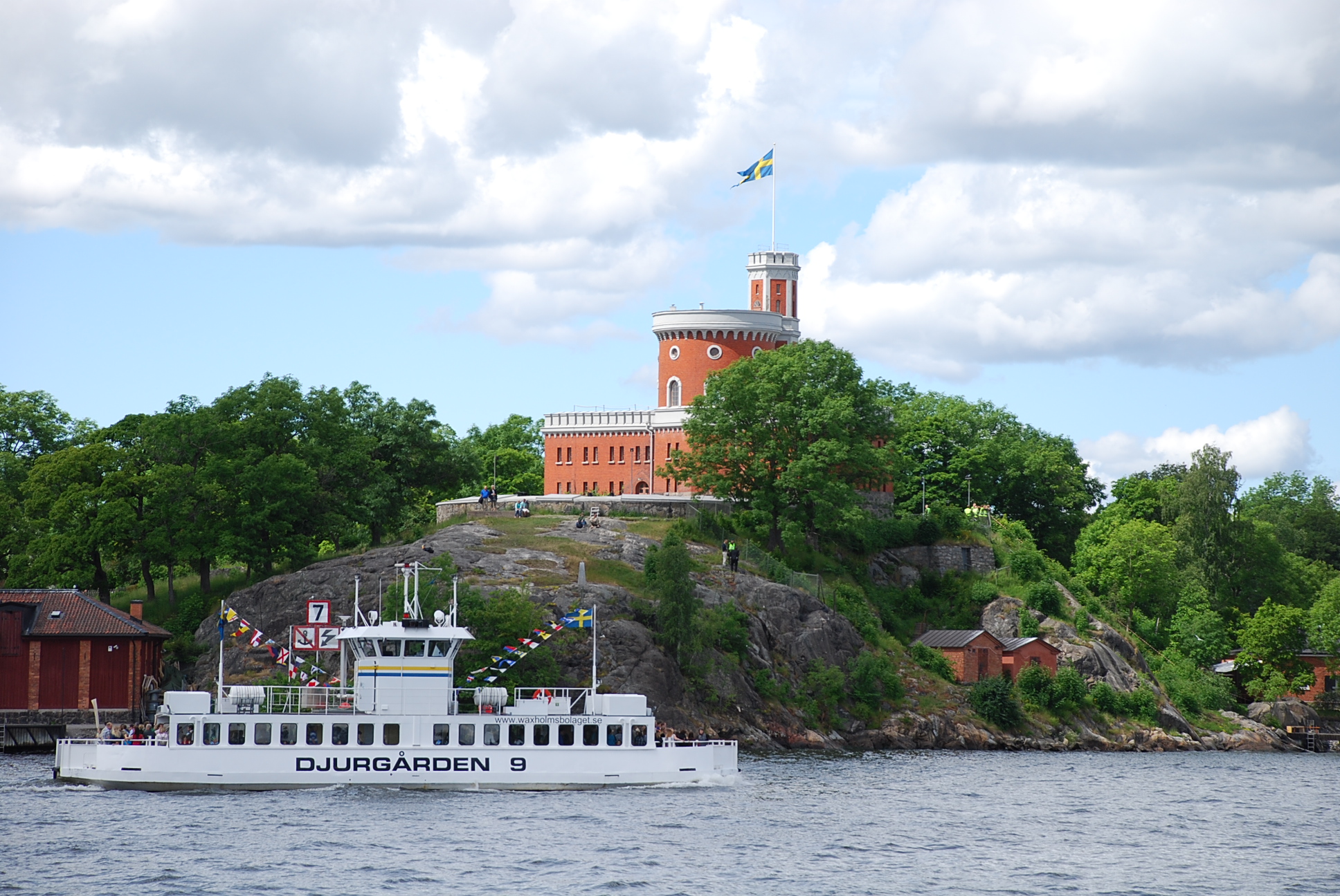Kastellet på Kastellholmen, omgivet av grönska. I förgrunden djurgårdsfärjan.