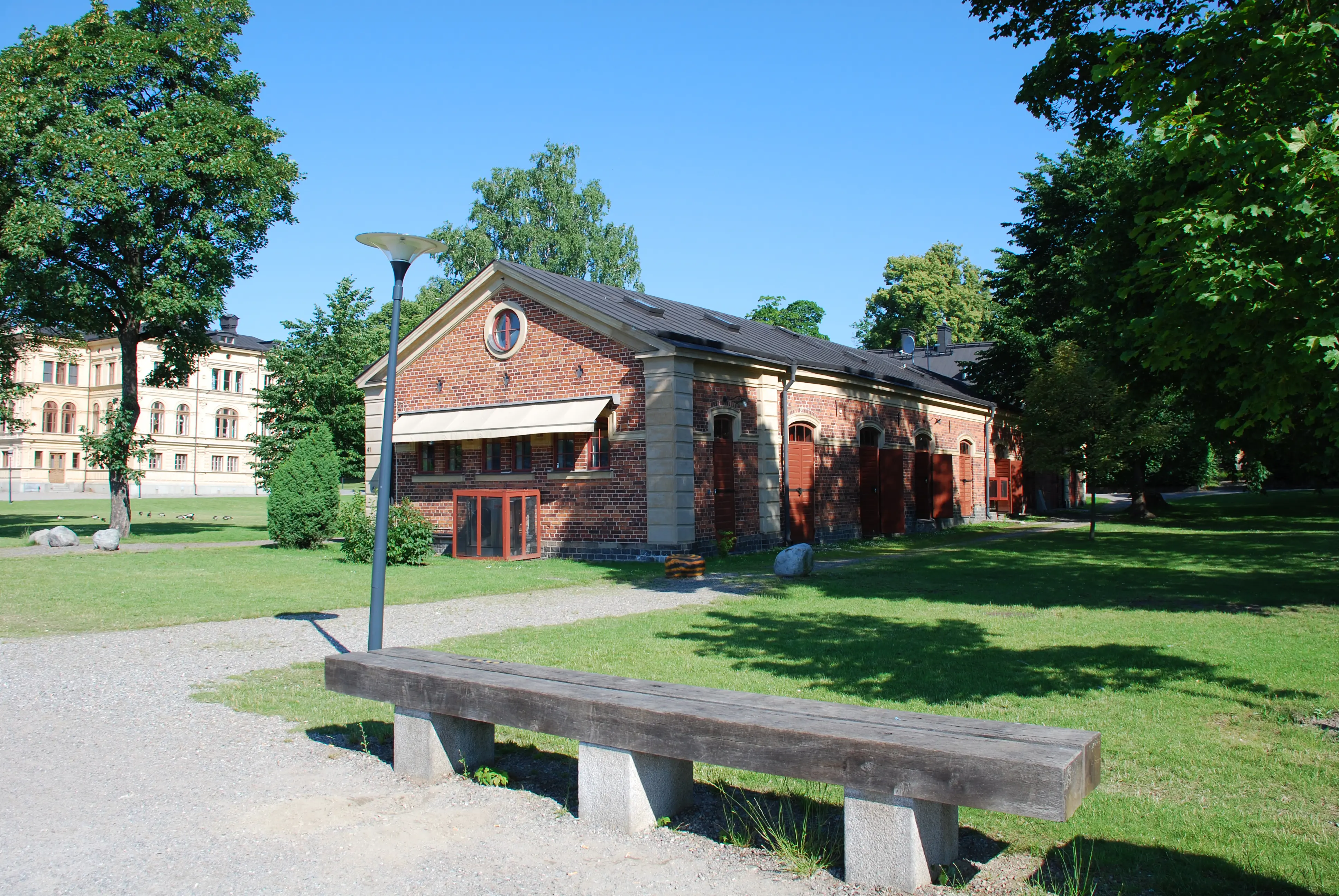 Västra uthuslängan, Skeppsholmen. Låg tegelbyggnad med klassicistisk dekor. Sommar och grönska.