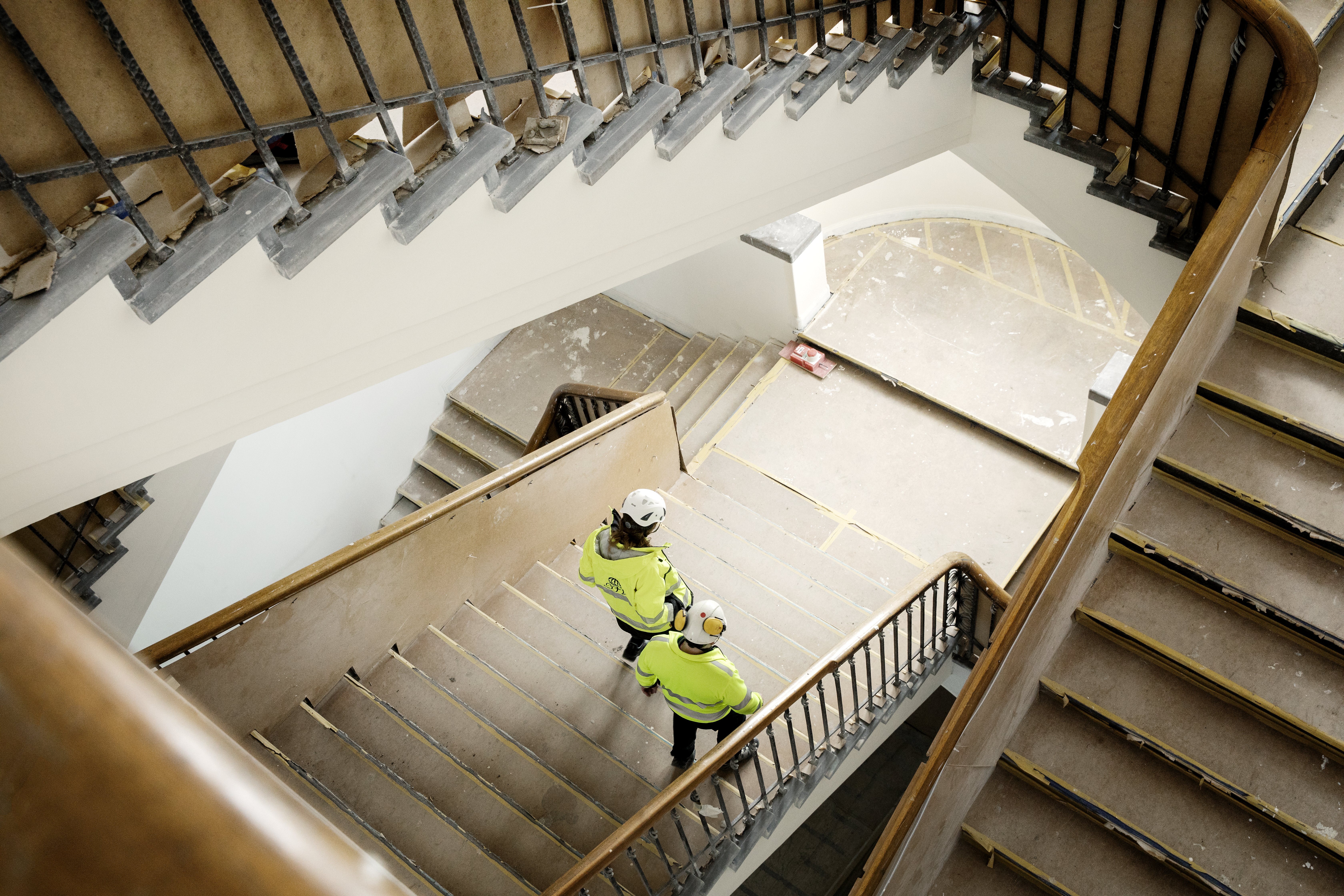 Två personer i varsel-kläder och hjälm sedda ovanifrån när de går nedför en trappa. Golv och trappor täcks av bygg-papp.