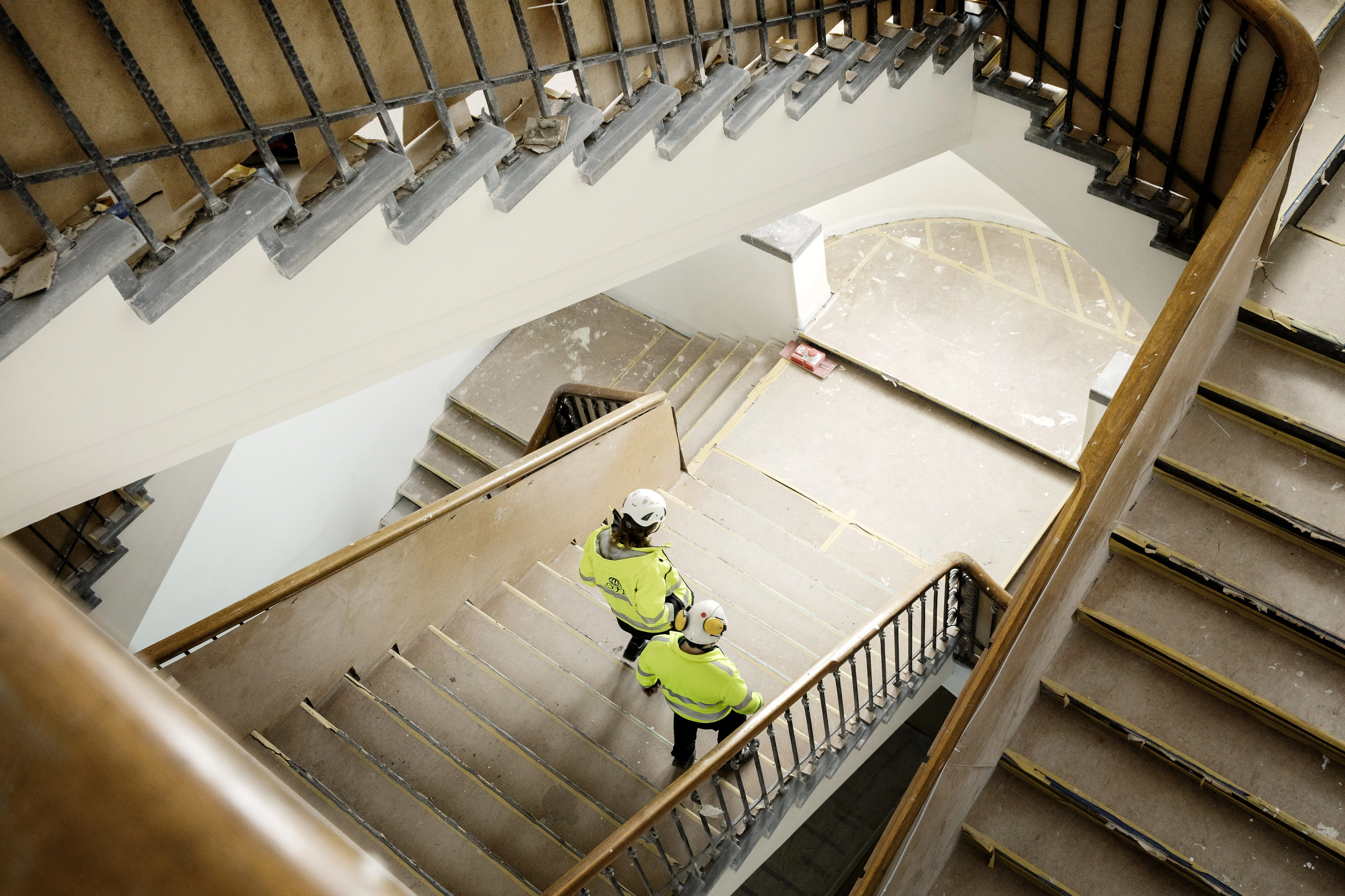 Två personer i varsel-kläder och hjälm sedda ovanifrån när de går nedför en trappa. Golv och trappor täcks av bygg-papp.