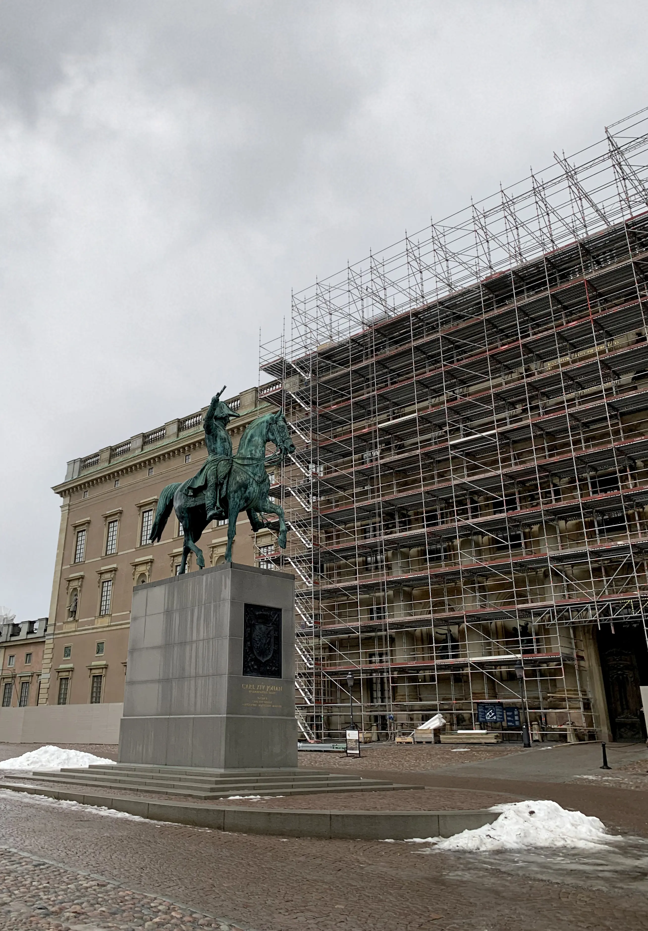 En stor byggställning till höger i bild, framför Stockholms slott som har en rosabrun rikt smyckad fasad. I förgrunden en staty föreställande Karl XIV Johan på en häst.