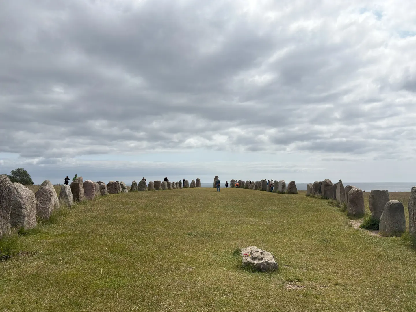 Ales stenar. Vy på längden genom skepps-sättningen mot havet. Några besökare står bland stenarna.