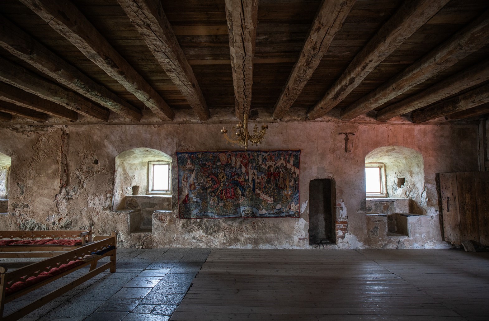 Stort rum med trägolv, träbjälkar i taket samt en stor och färggrann väggtextil på en av väggarna.