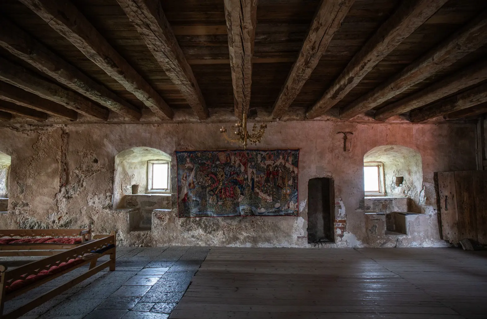 Stort rum med trägolv, träbjälkar i taket samt en stor och färggrann väggtextil på en av väggarna.