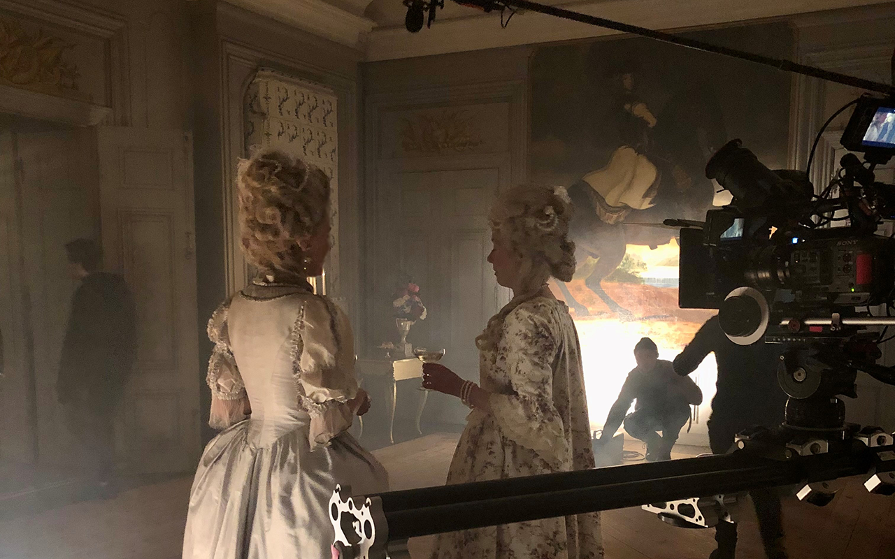 Två kvinnor i rokoko-kläder står bland filmutrustning i en sjuttonhundratals-salong.