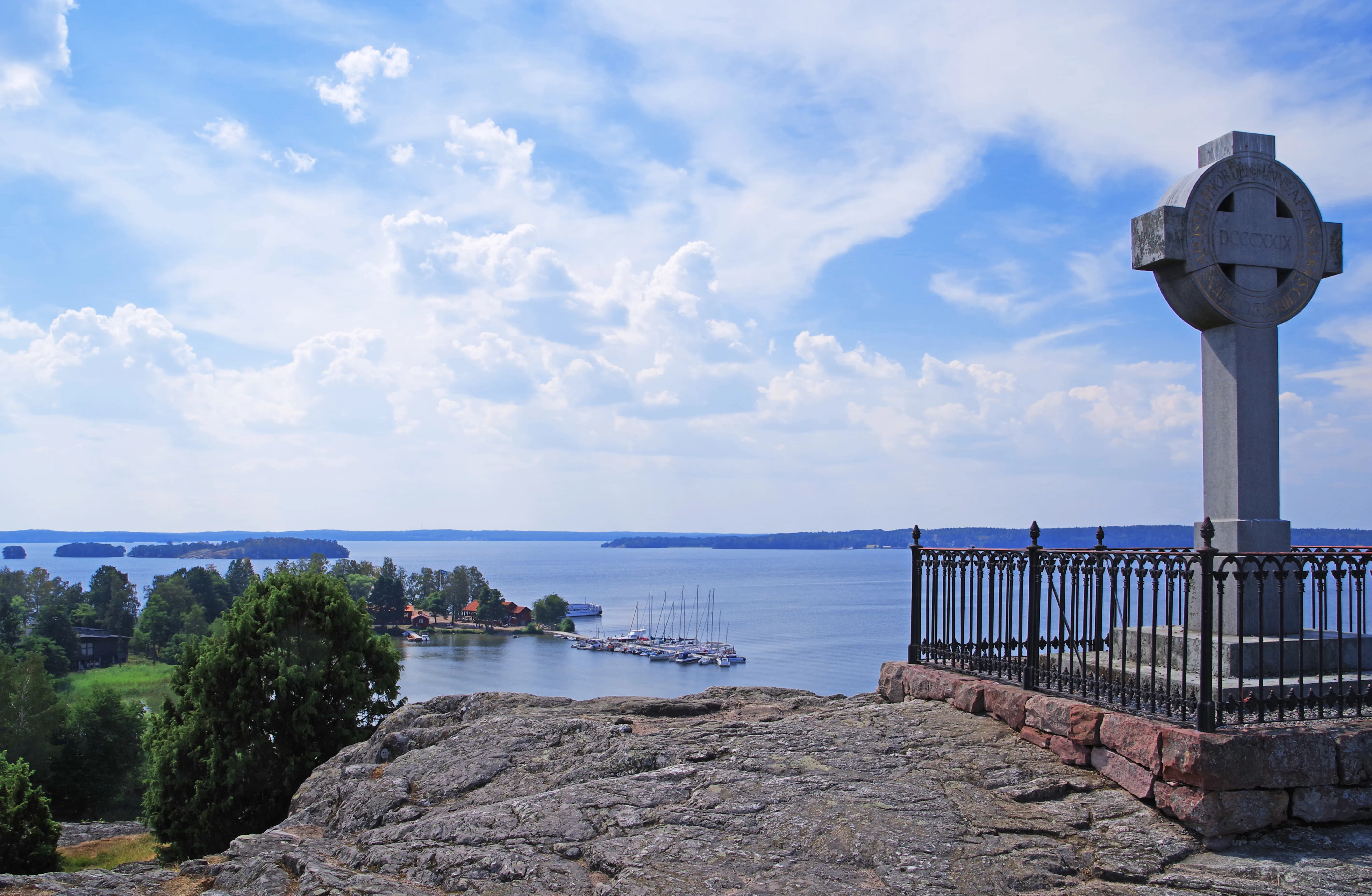 Ansgarskorset på Björkö. Ett stenkors omgärdat av ett gjutjärns-staket på en klippa. Utsikt över museet och Björköfjärden.