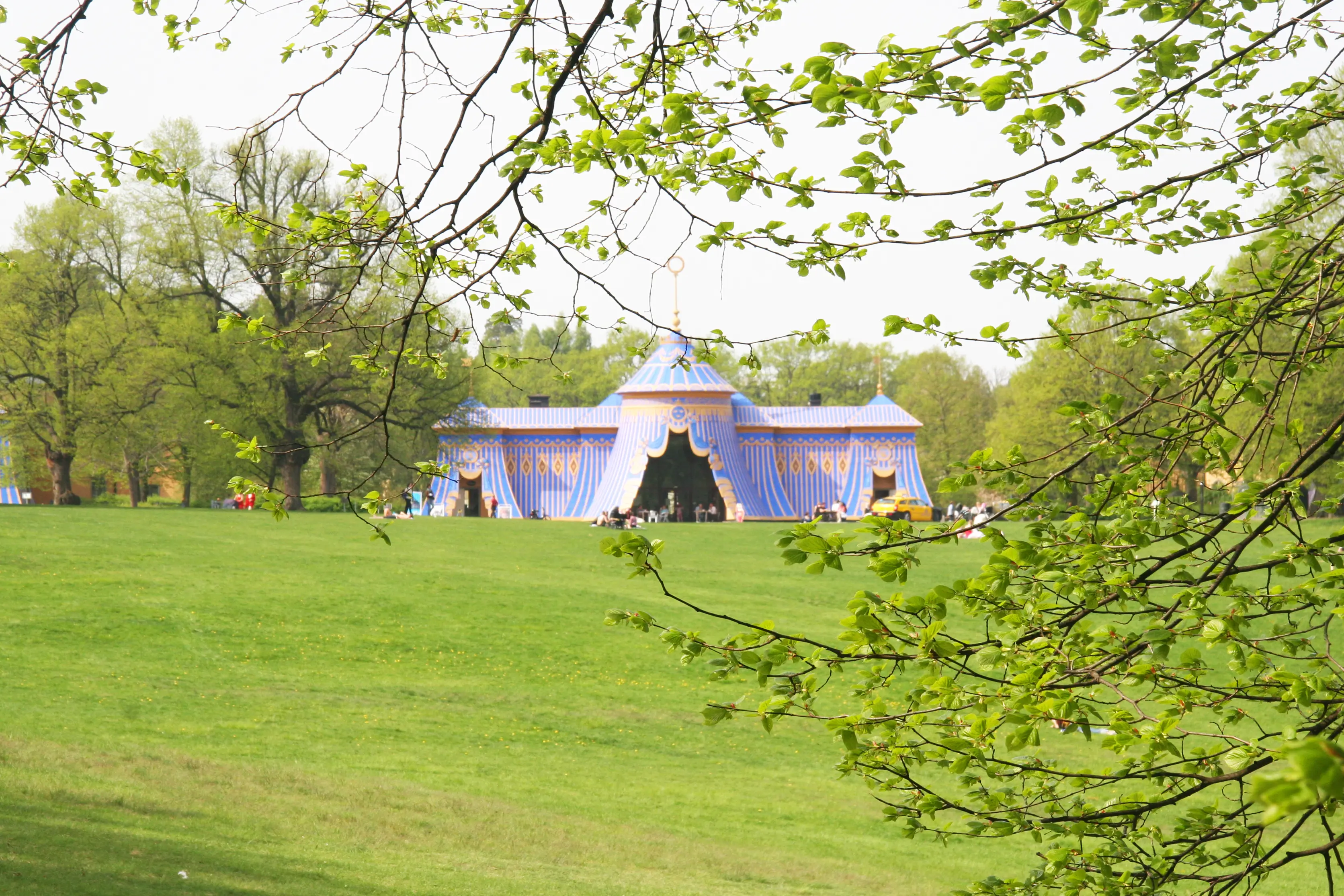 Koppartälten i Hagaparken, sedda genom spirande löv-verk. Många besökare fikar eller promenerar utanför byggnaderna.