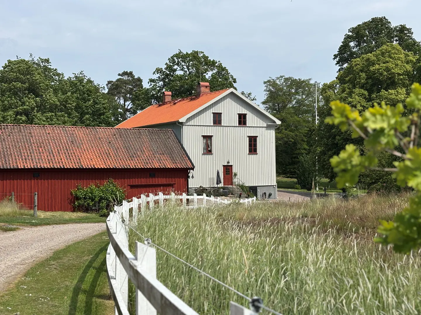 En grusväg längs med ett staket som leder mot en röd förrådsbyggnad och det ljusgrå bostadshuset.