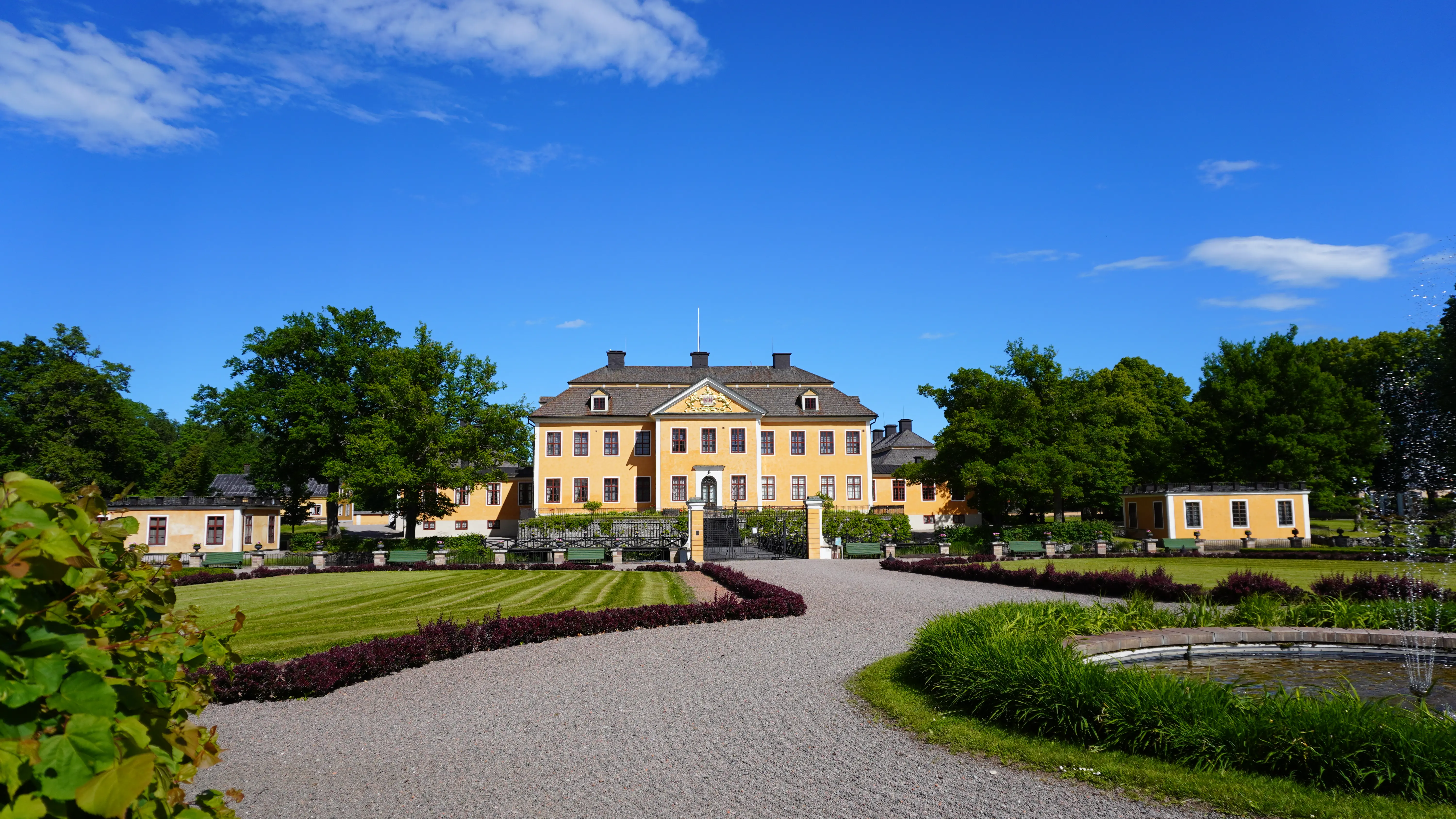 Lövsta bruks herrgård sedd från barockträdgården. Huvudbyggnaden och de små sidobyggnaderna lyser gula bland lummiga, grönskande träd.