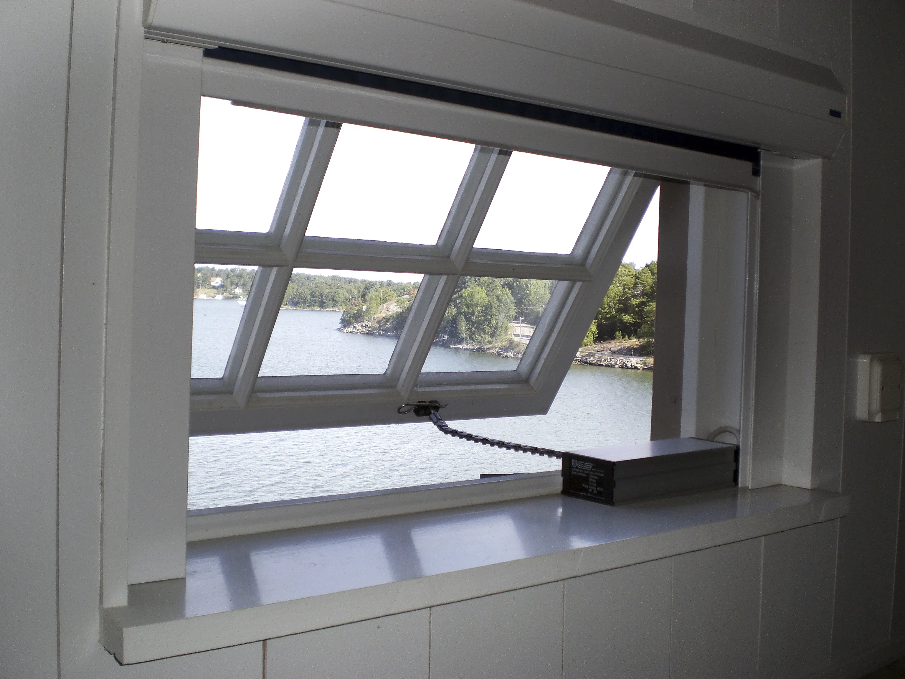 Närbild på en vit fönsterkarm och fönsterbräda med ett avlångt spröjsat fönster på glänt. Utsikt över somrig skärgårdsmiljö.