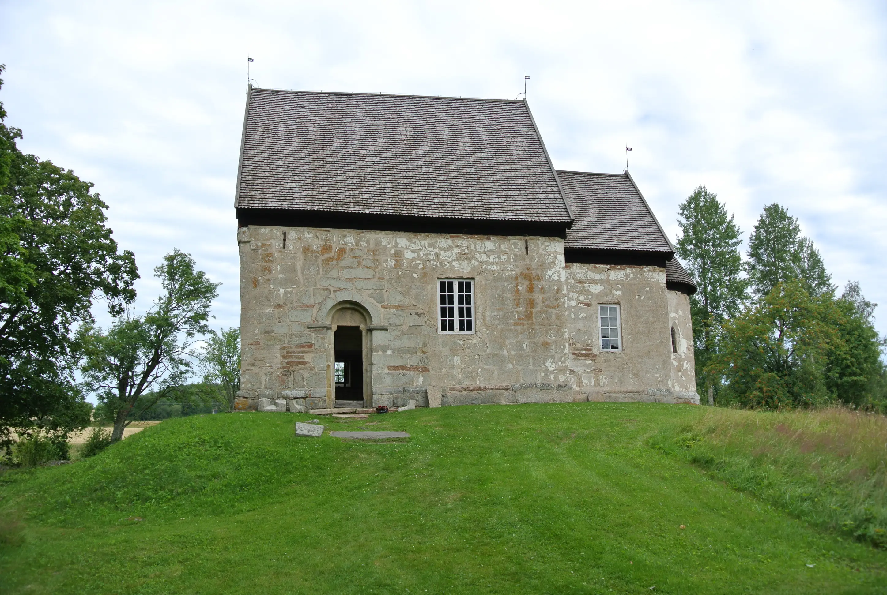 Suntaks gamla kyrka. Enkel stenkyrka med spåntak. Sommar och grönska.