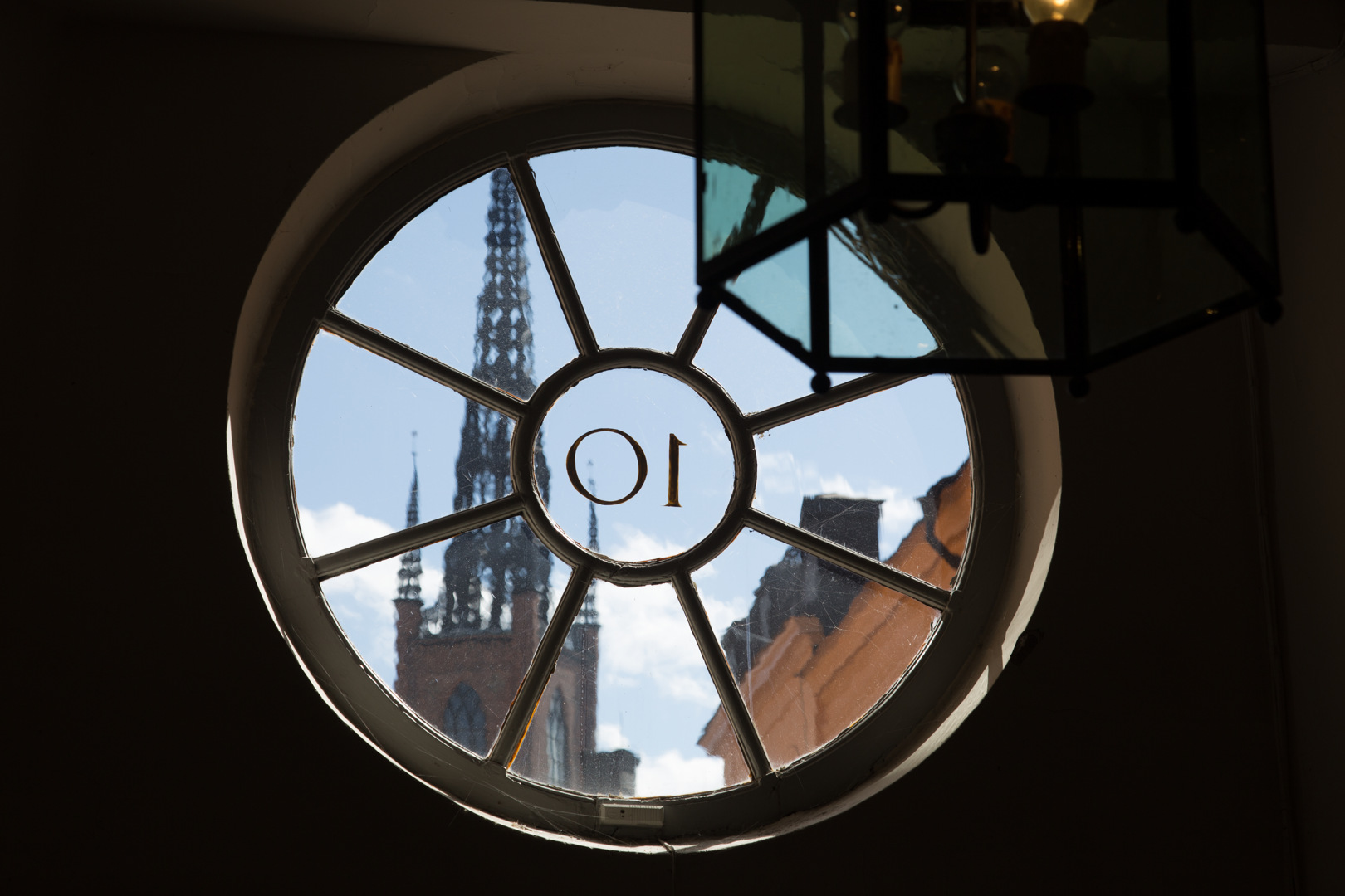 Rosenhaneska. Inredningsdetalj. Runt, spröjsat fönster ovanför porten. Genom glaset syns Riddarholmskyrkans spira.