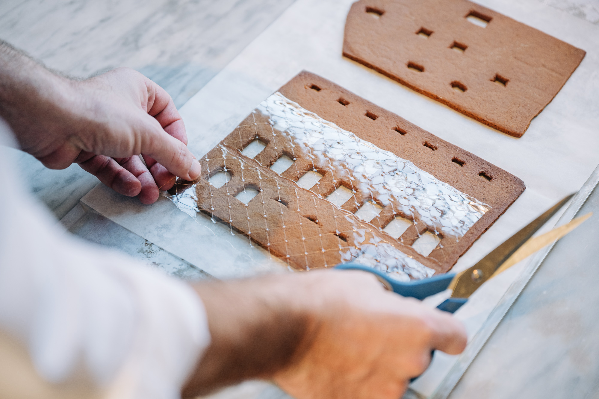 Ett par händer måttar ett ark med gelatinblad till fönsterna på en gräddad del till ett pepparkakshus.