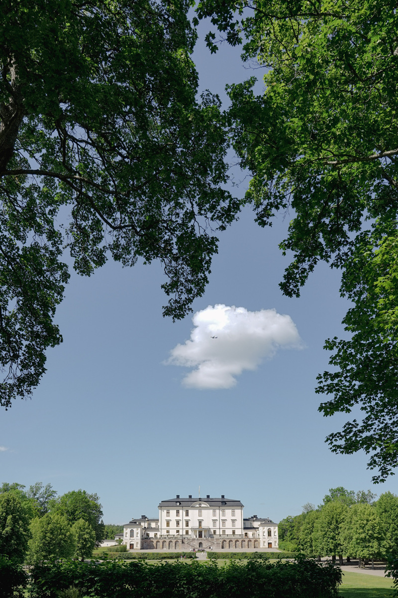 Rosersbergs slottspark. Höga, lummiga träd ramar in vyn mot slottet över den stora gräsmattan.
