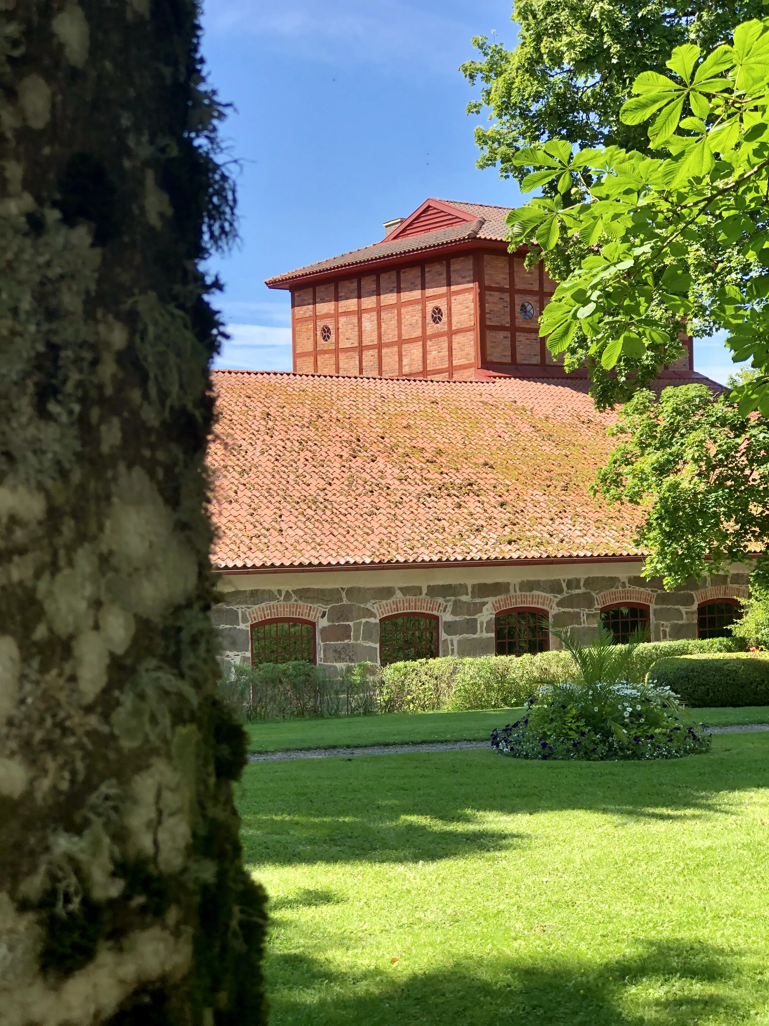 Gjuteriet. En låg, lång stenbyggnad med tegeltak. Bakom den syns masugnen, en hög korsvirkes-byggnad i tegel. Sommar och grönska.