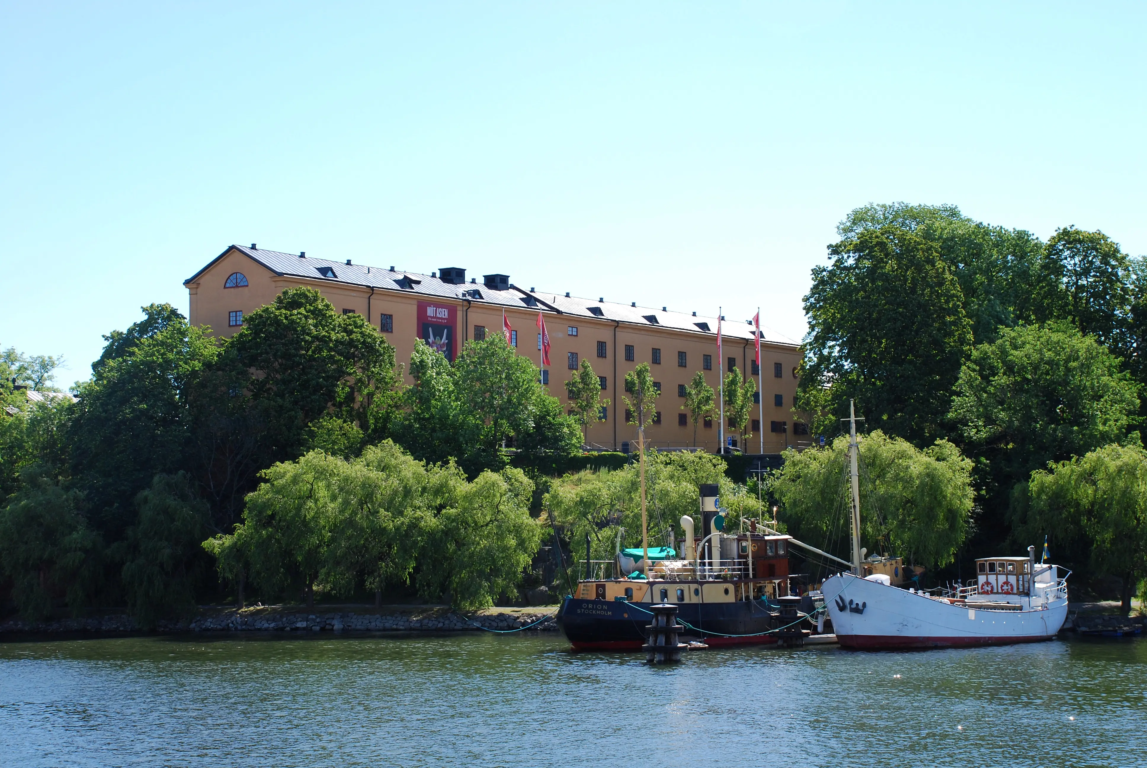 Östasiatiska museet sett över vattnet från Blasieholmen. En långsmal byggnad i tre våningar på en liten höjd. Sommar och grönska.