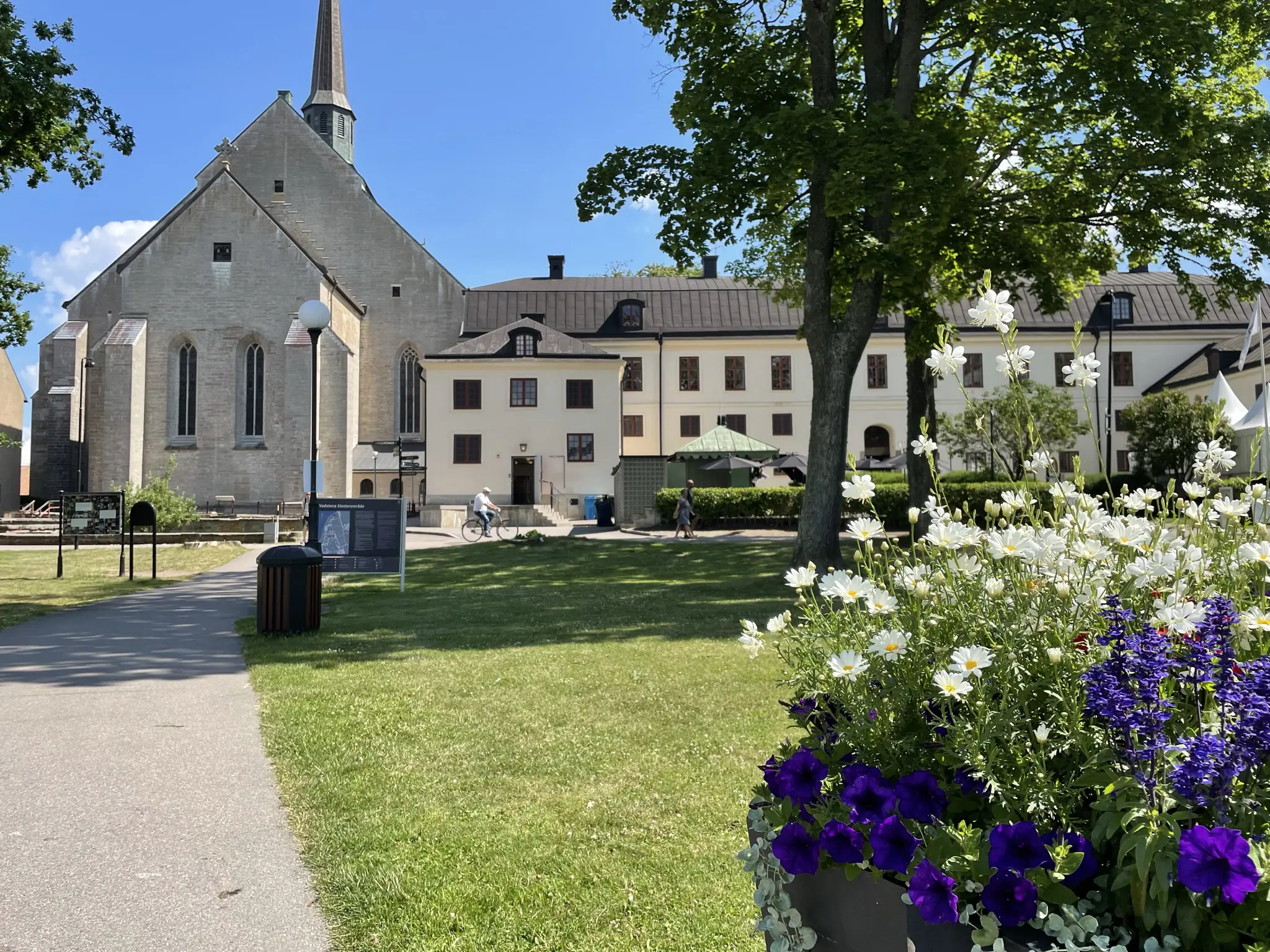 Munk-klostret i Vadstena. En vit trevåningsbyggnad, med en lägre flygel. Till vänster i bild syns klosterkyrkan. Sommar och grönska.
