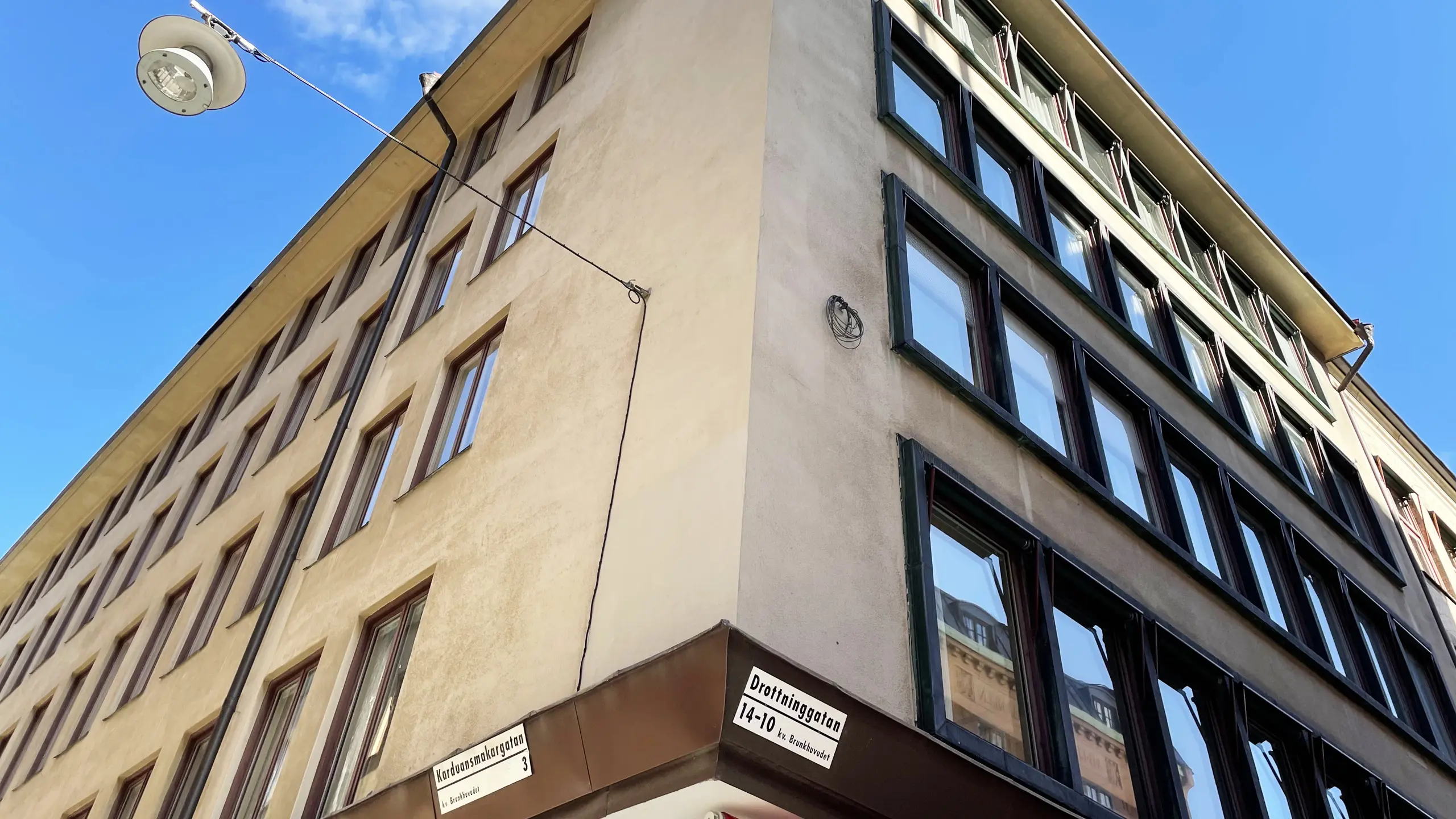 Hörnet på en byggnad med beigeputsad fasad och tätt sittande fönster. Gatuskyltarna säger att det är korsningen Karduansmakargatan-Drottninggatan.