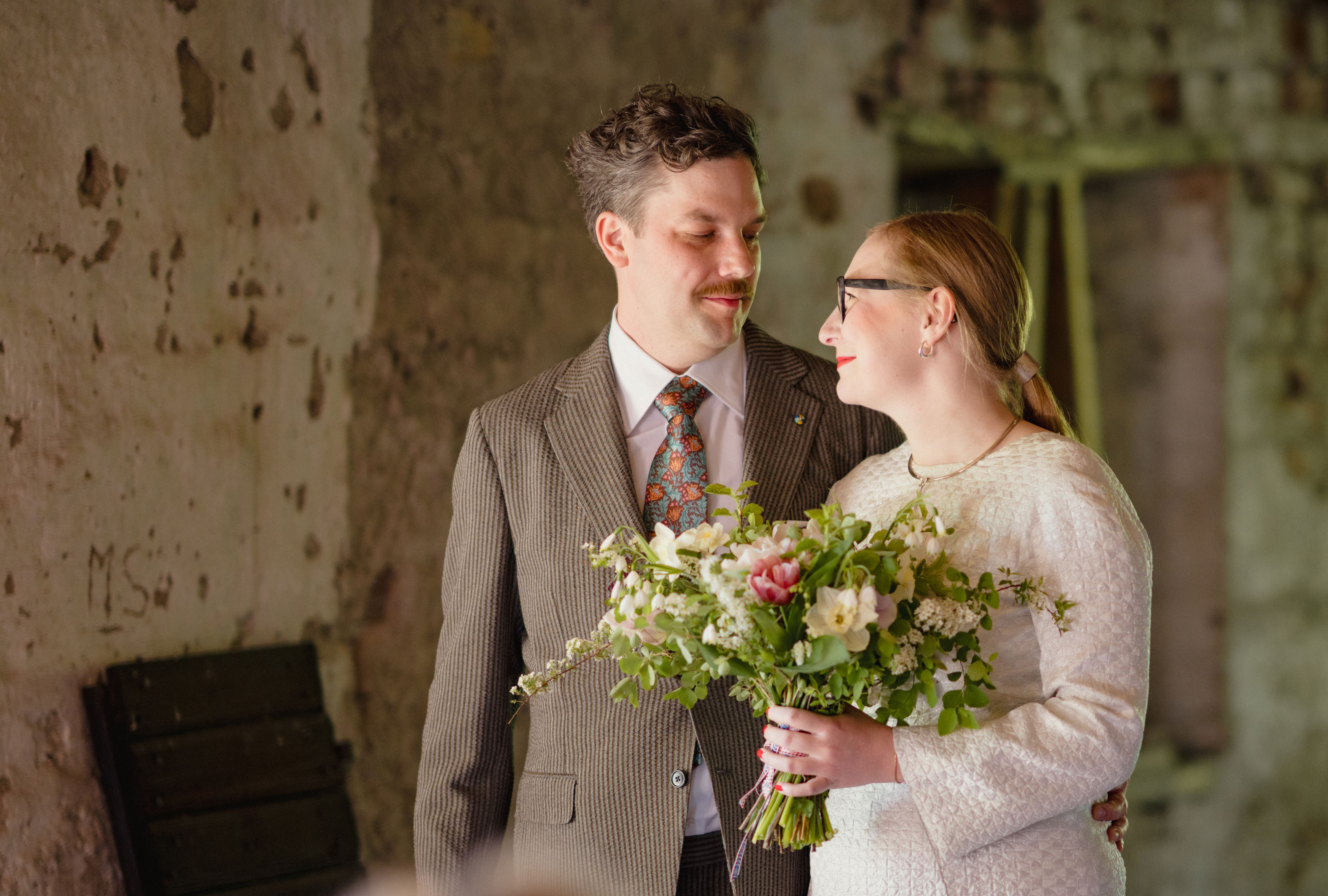 Vigsel mellan man i beige kostym och kvinna i vit klänning med blombukett
