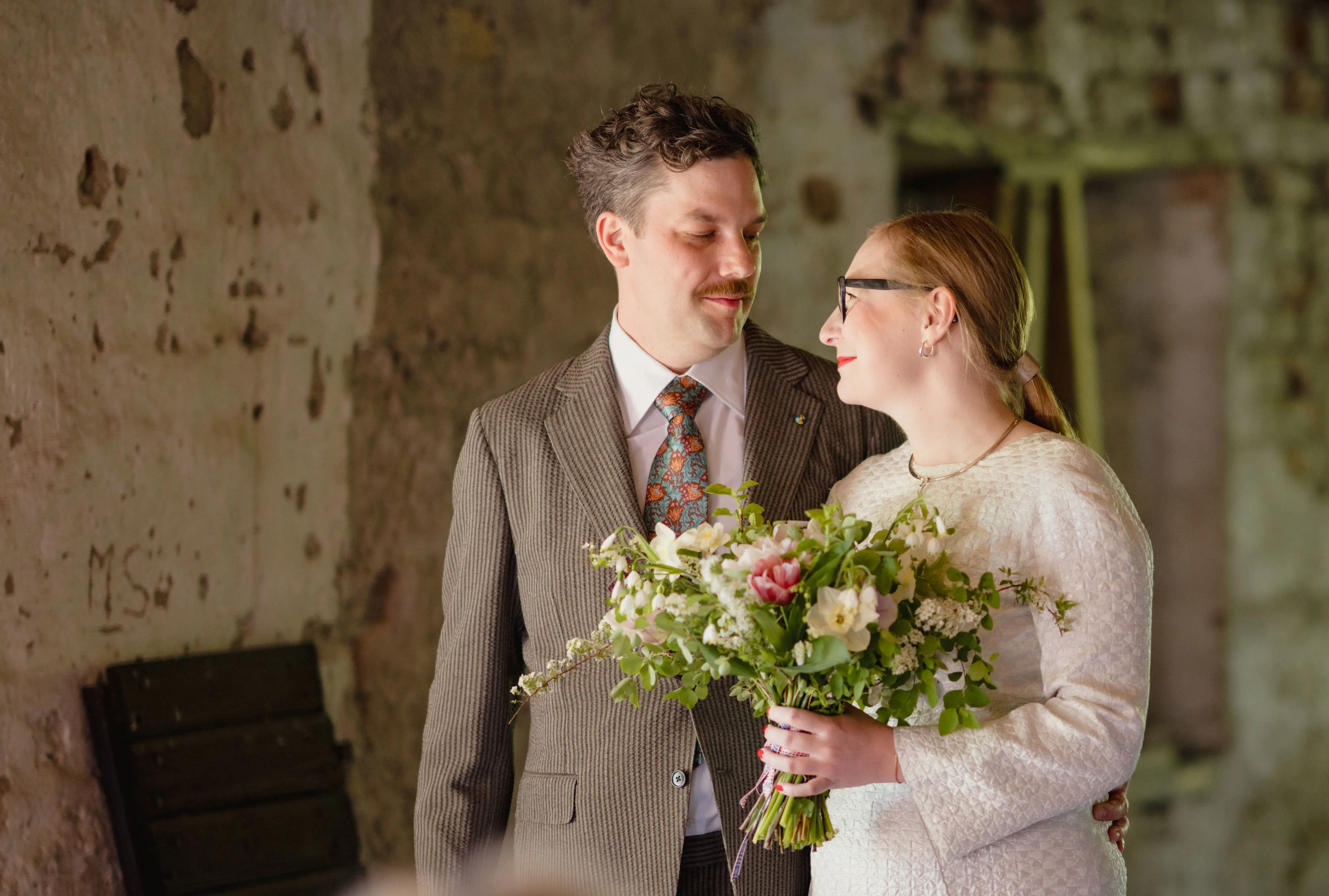 Vigsel mellan man i beige kostym och kvinna i vit klänning med blombukett