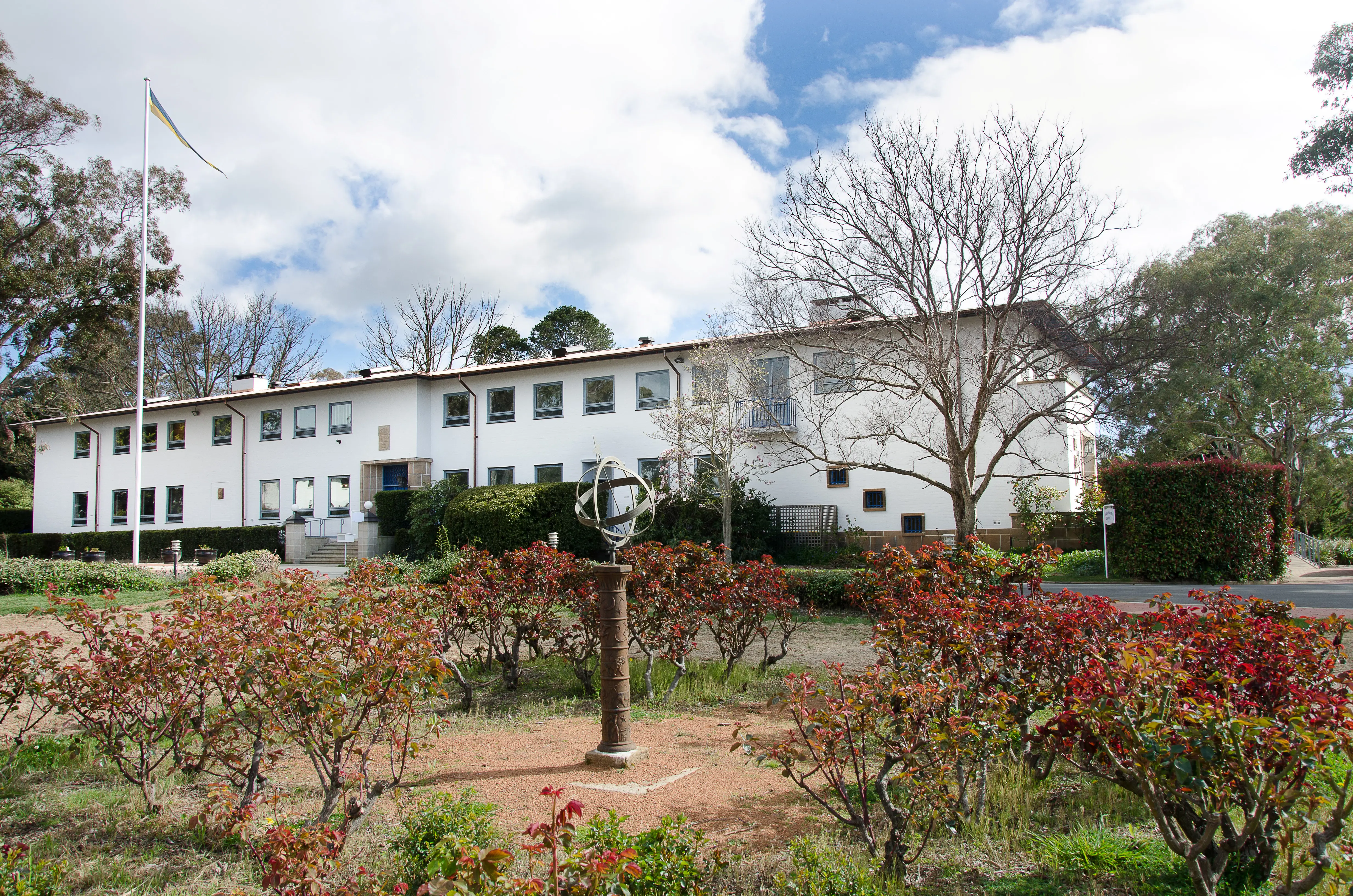 Ambassaden i Canberra. En vitputsad, funktionalistisk tvåvåningsbyggnad. Framför byggnaden är en trädgård med ett solur.