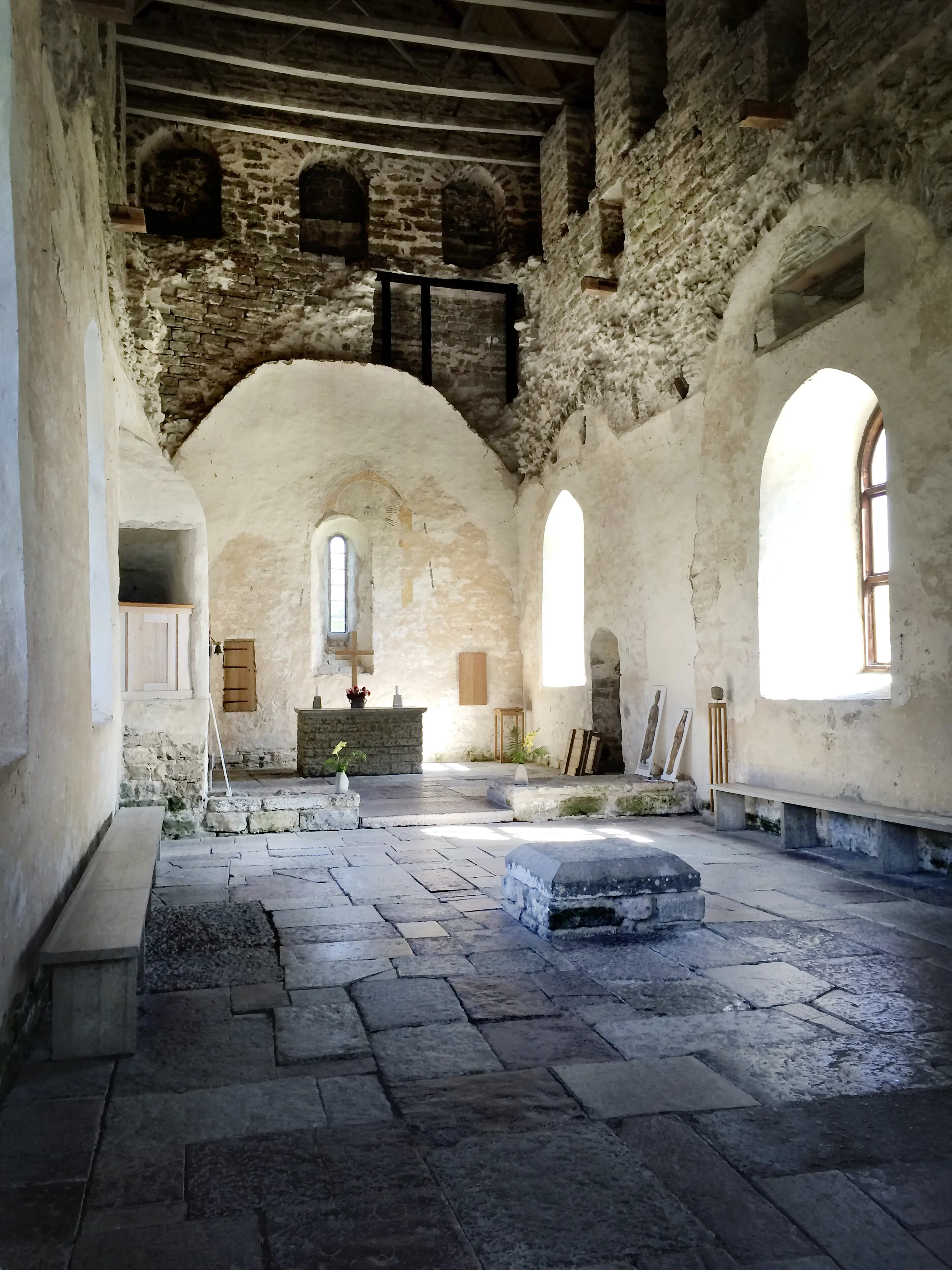 Ett kyrkorum med mörkt stengolv, ljust putsade väggar och högt i tak. Längst fram i kyrkan syns altaret byggt i mörka stenar. På altaret står ett kors i ljust trämaterial.