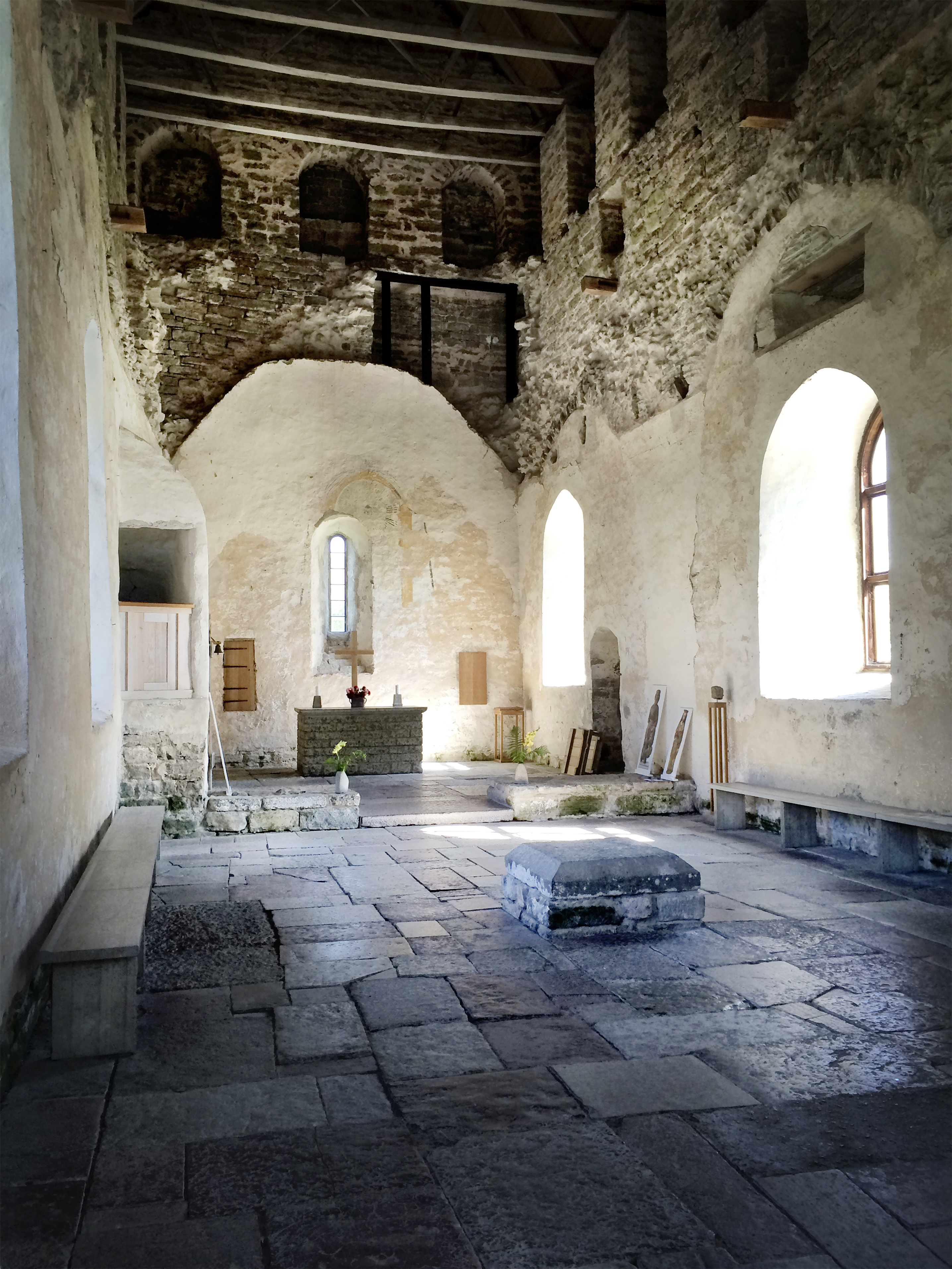 Ett kyrkorum med mörkt stengolv, ljust putsade väggar och högt i tak. Längst fram i kyrkan syns altaret byggt i mörka stenar. På altaret står ett kors i ljust trämaterial.