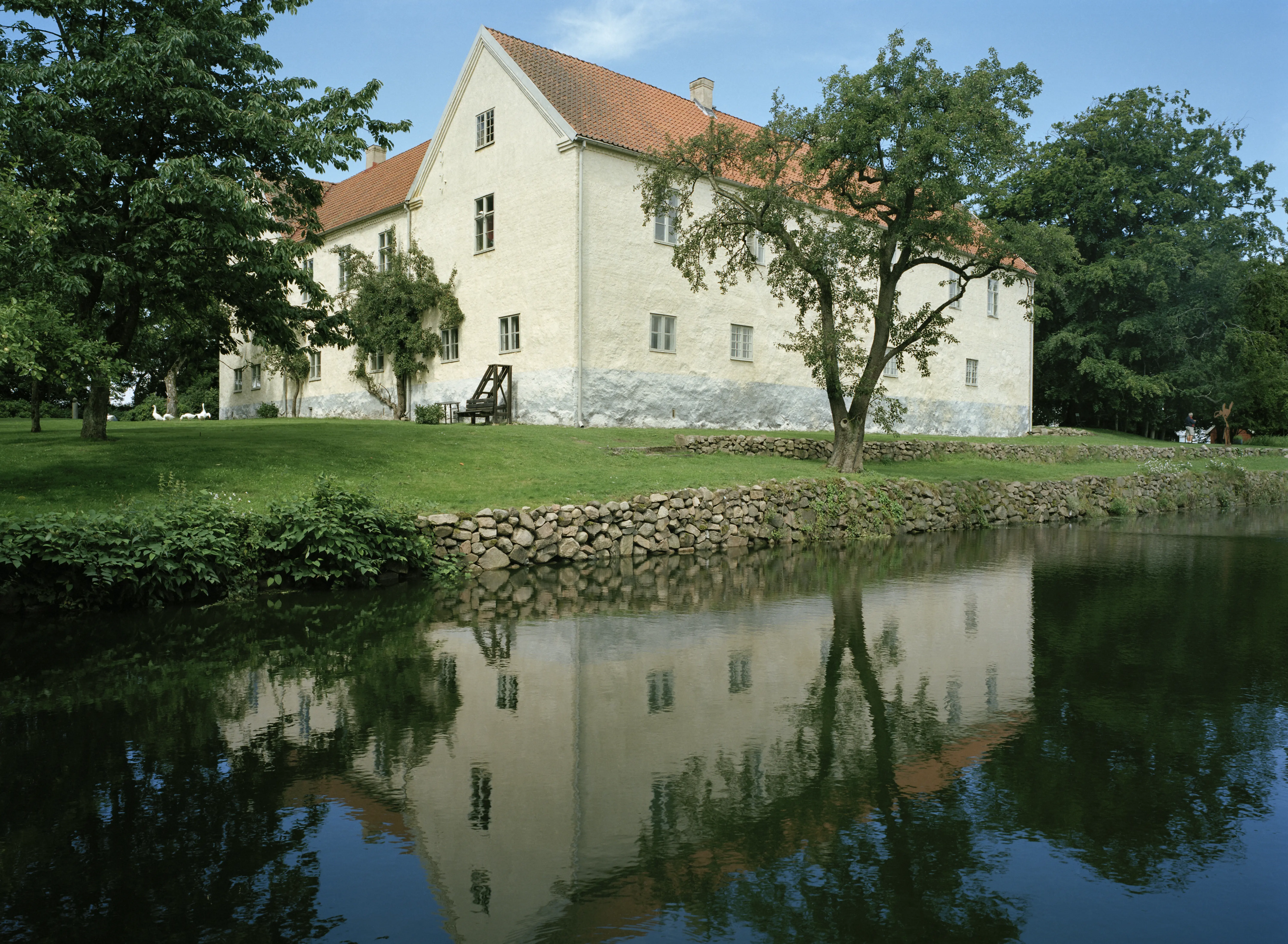Tomarps kungsgård. Östra längan, en vitputsad stenbyggnad, speglas i en damm. Sommar och grönska.