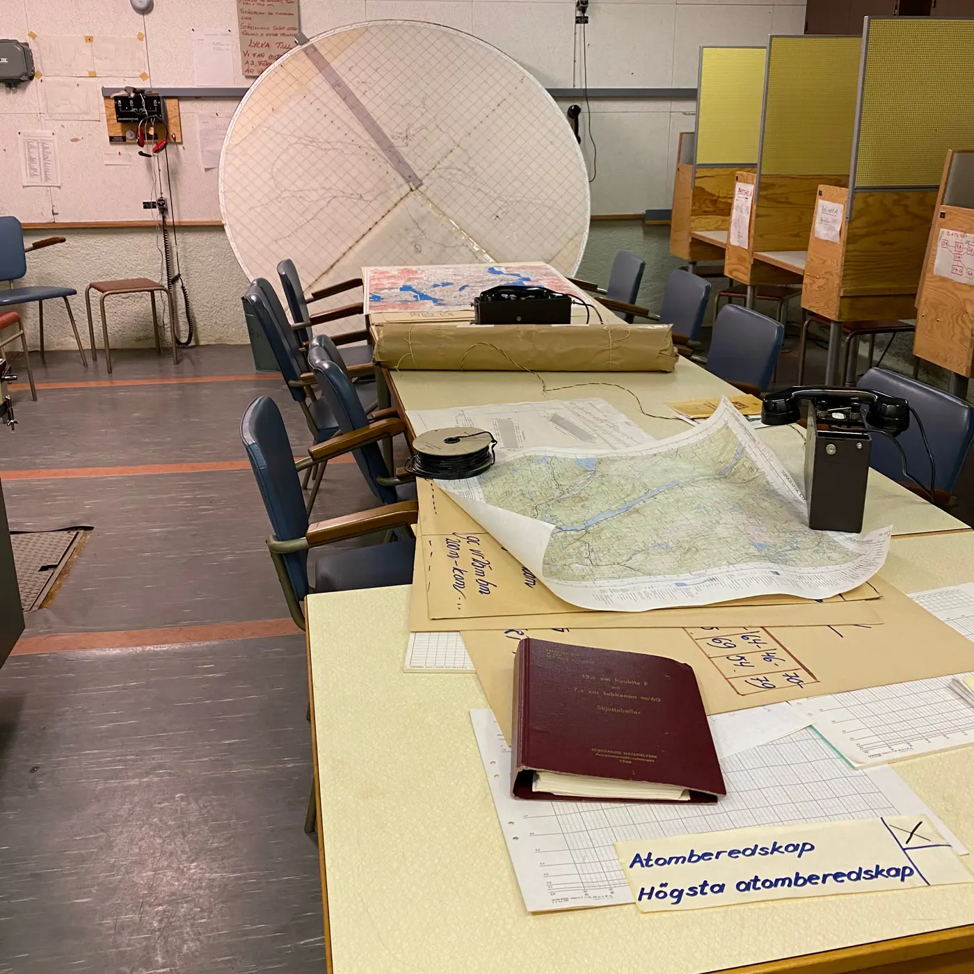 Ett rum med vita väggar med djud-dämpande material. Grå linoleummatta på golvet. Ett bord är fyllt med papper och kartor. På en lapp står "Atomberedskap, Högsta atomberedskap". Till höger i bild syns en rad med bås med arbetsplatser.