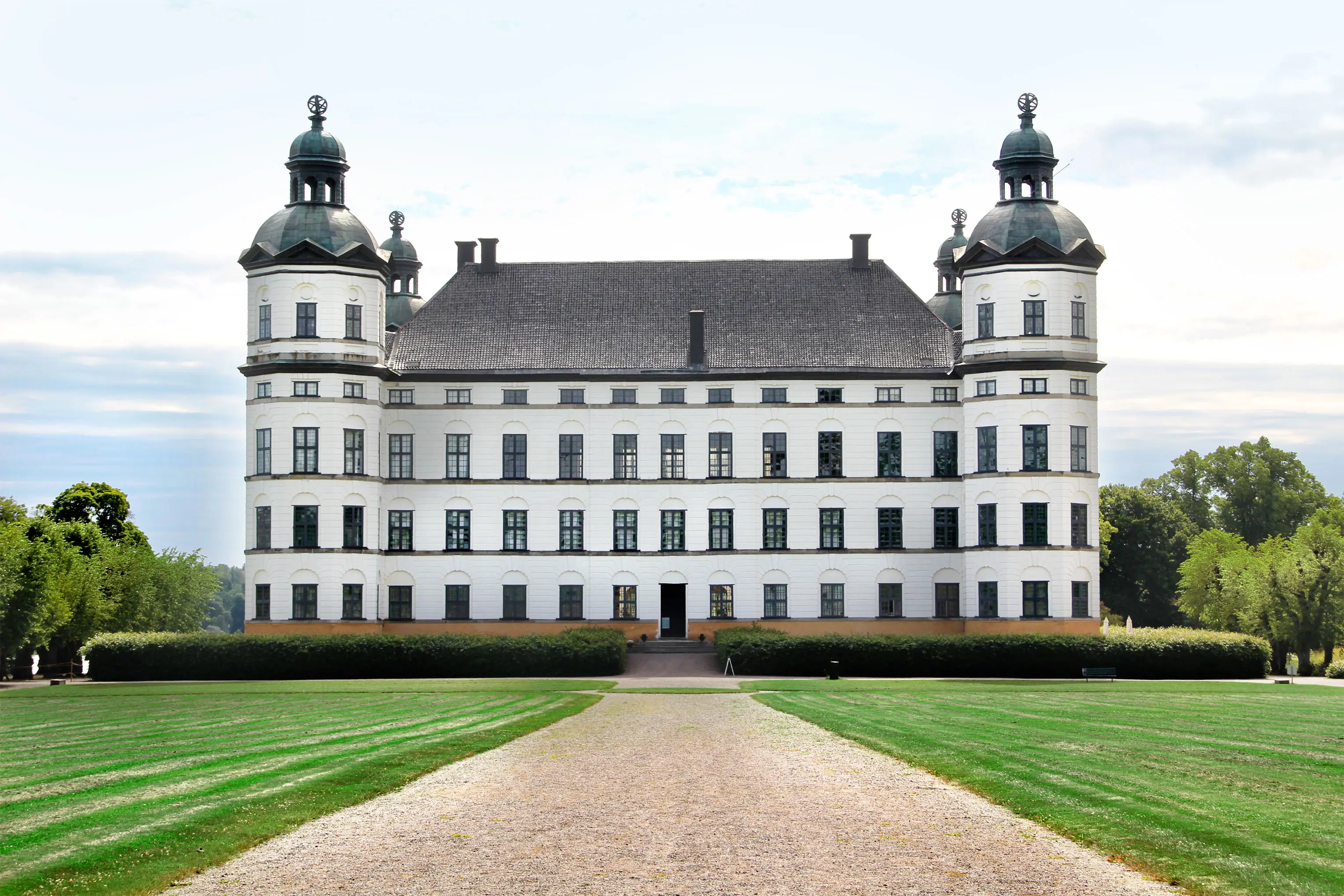 Vitt slott med fyra torn omgiven av gräsmatta, grusgångar och träd.