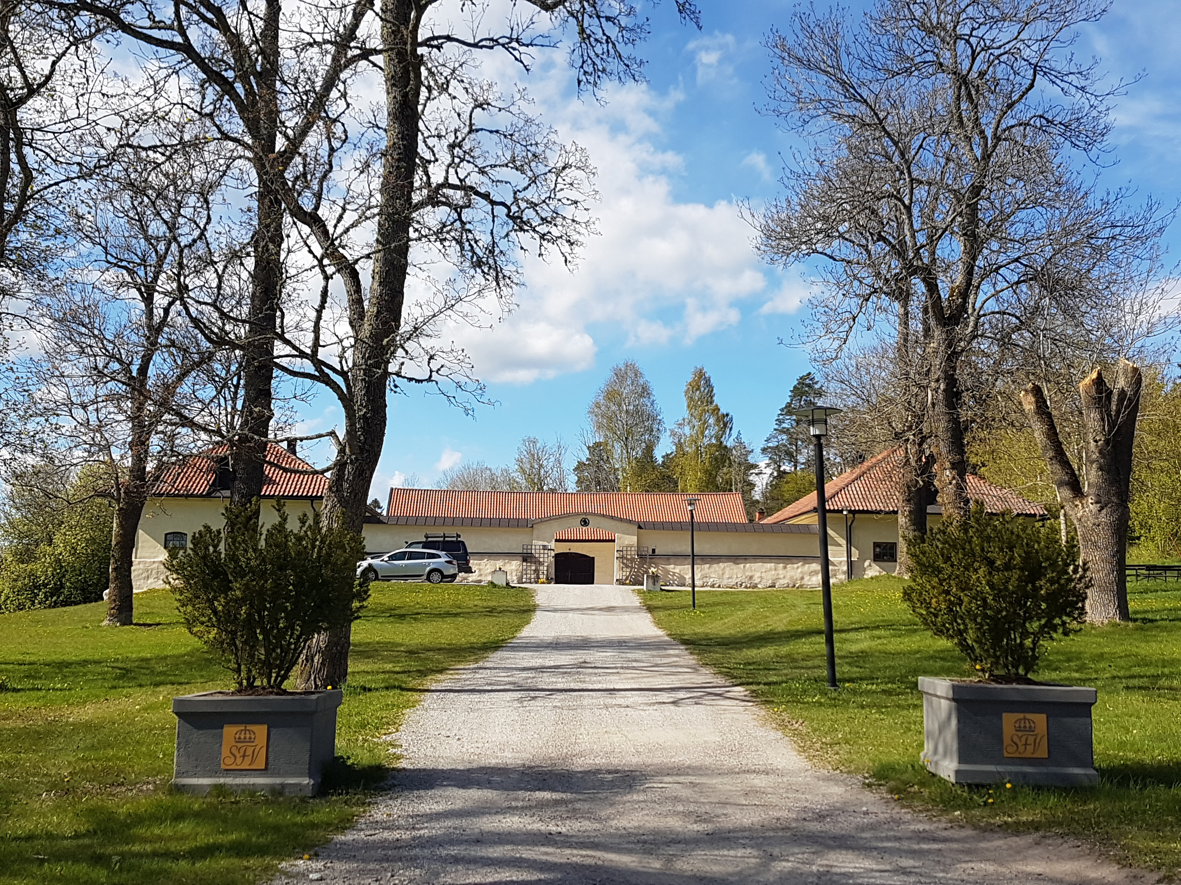 Rosersbergs kungsgård. Före detta ox-stall. Tre putsade stenbyggnader, sammanbyggda av murar. Vår och grönska.