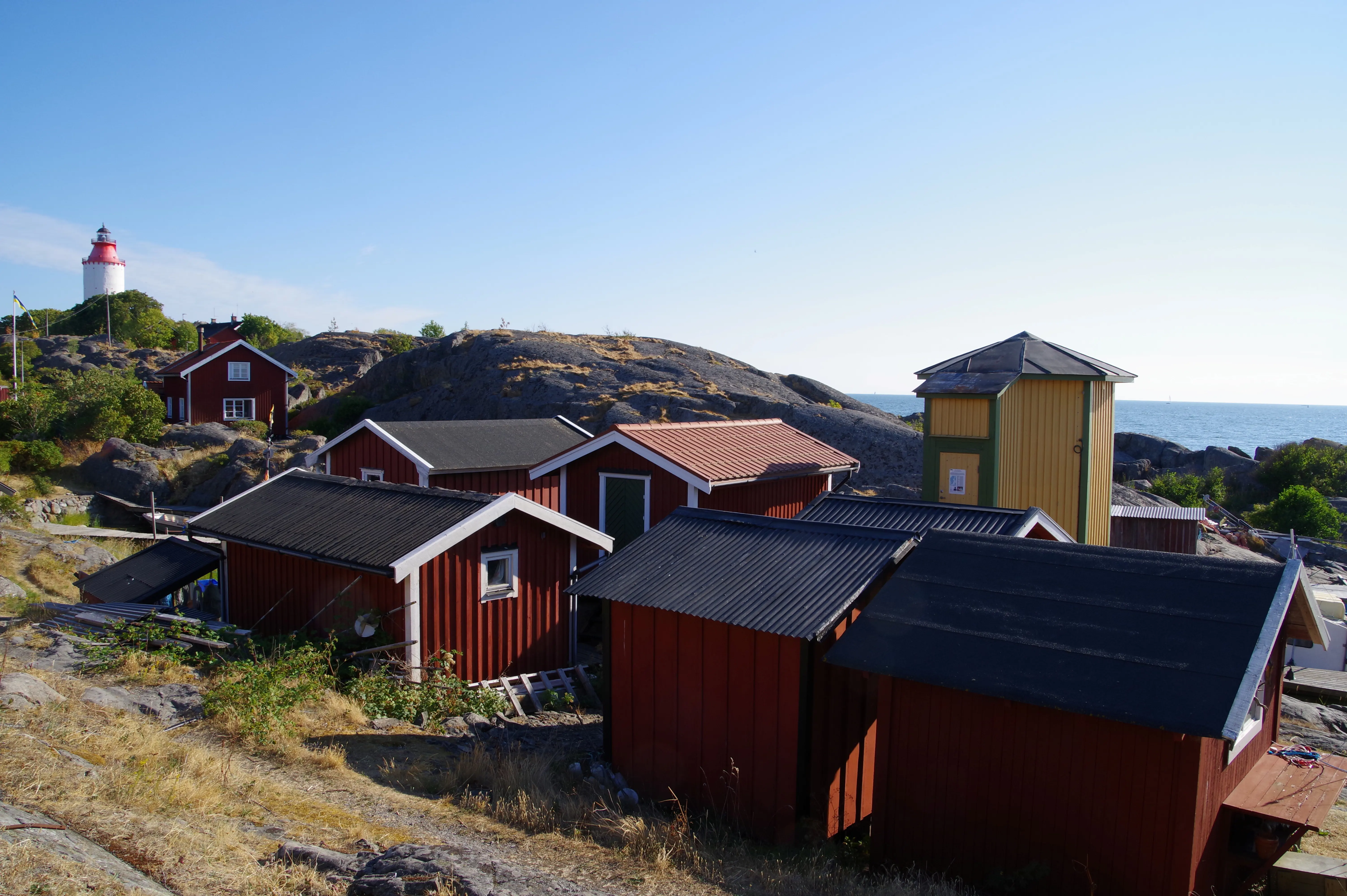 Träbebyggelse bland klipporna på Öja. Små, röda stugor och en lite högre gul byggnad. Långt till vänster i bild skymtar fyren.