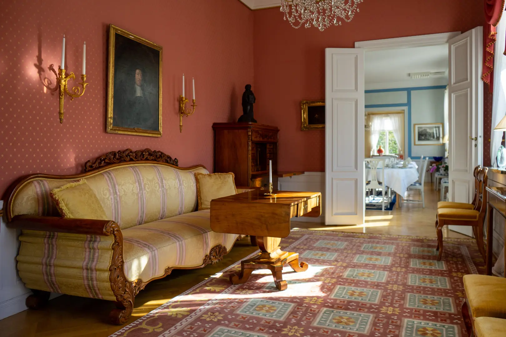 Interiör med röda tapeter, ett inramat porträtt och en soffa med gul klädsel.