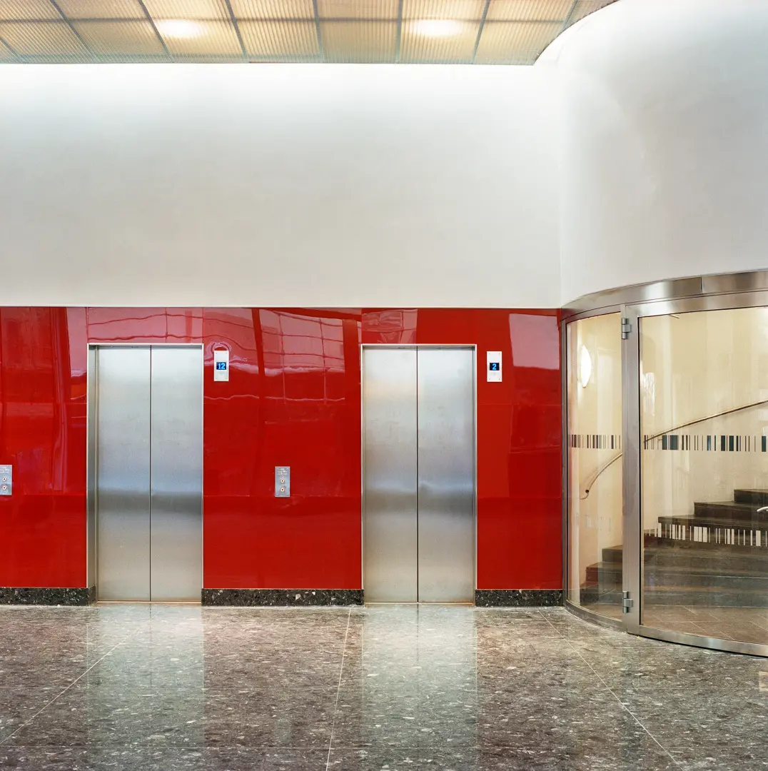 Blank röd vägg med två hissdörrar av metall. Till höger syns ett trapphus omslutet av en rundad glasyta.