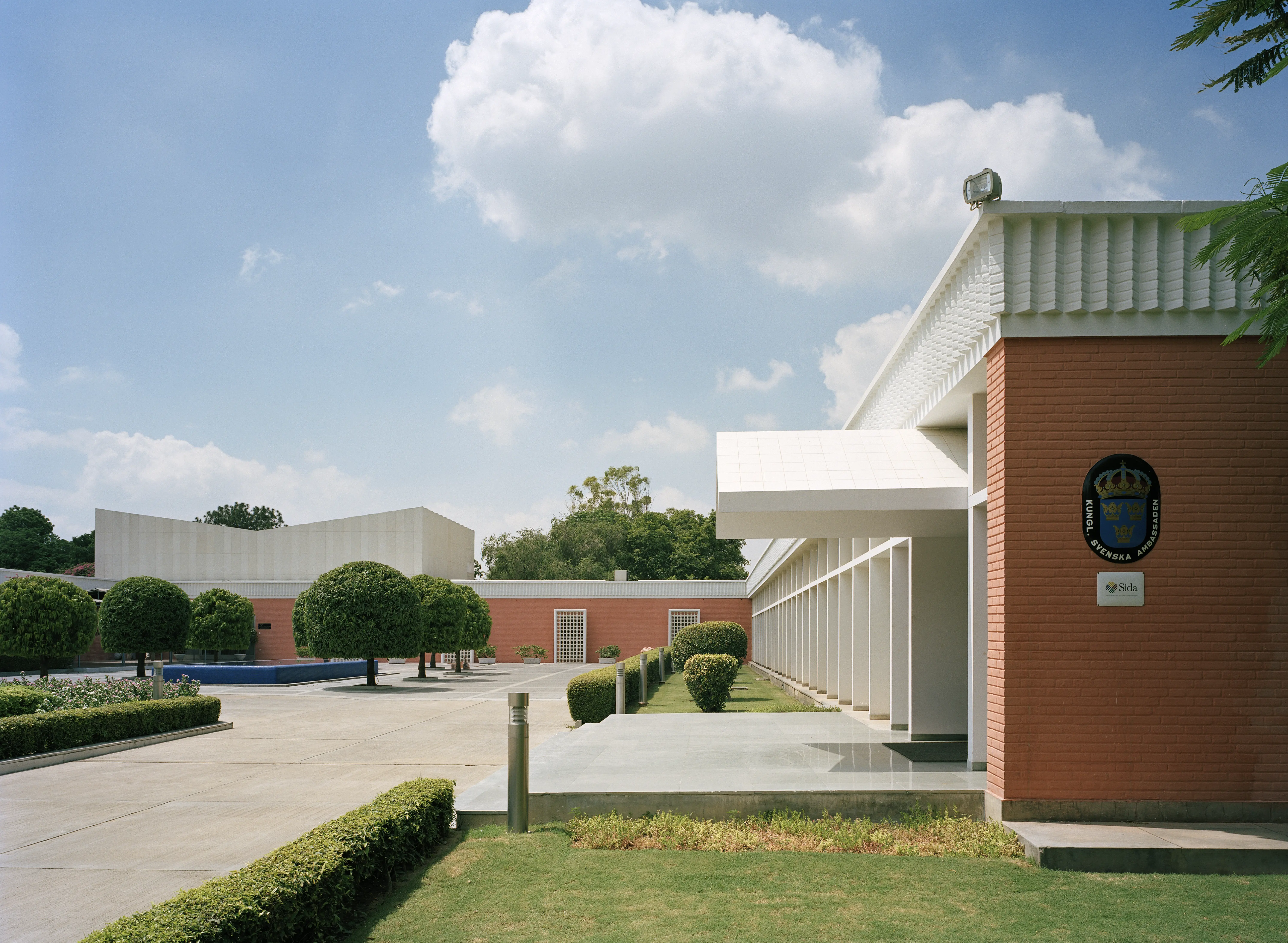 Ambassaden i New Delhi. Låga, långsträckta byggnader i rosa och vitt tegel omgärdar en gård med fontän och planteringar.