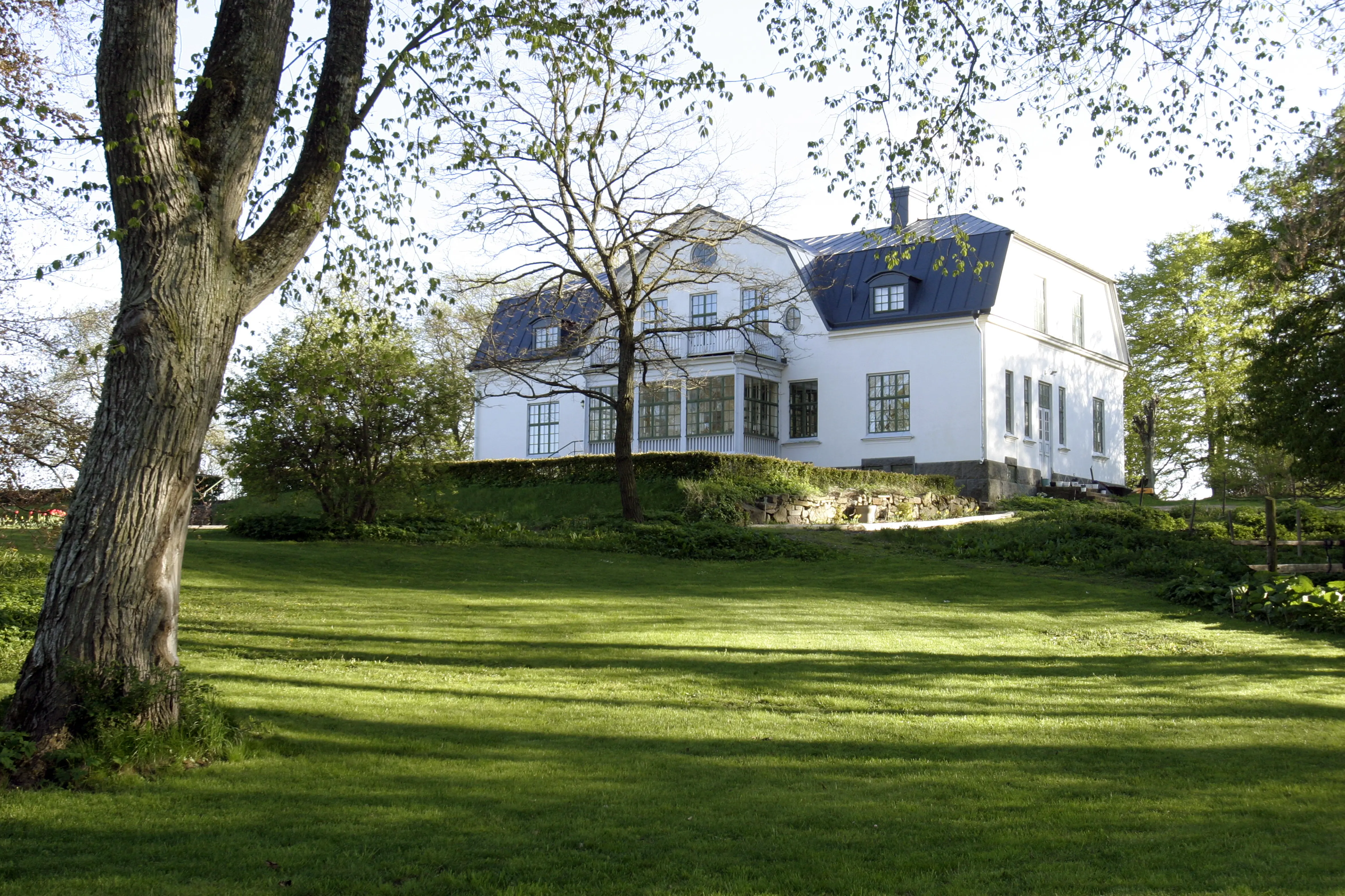 Mangårds-byggnaden på Alvastra gård. En vitputsad tvåvånings-byggnad med glasveranda och balkong. Vår och skir grönska.