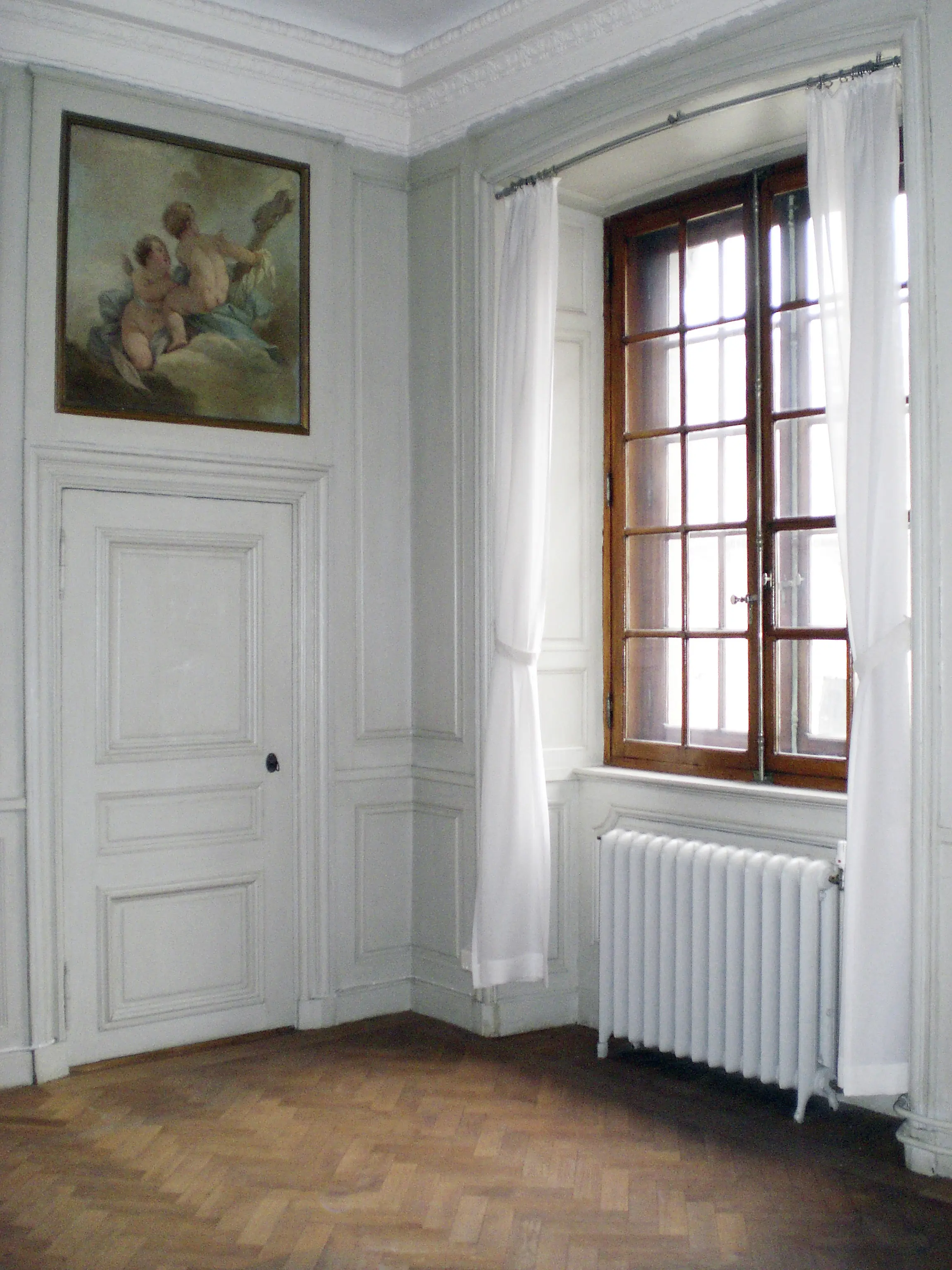 Ett hörn av en gammal byggnad med högt i tak och stuckaturer. Till vänster en dörr med en tavla ovanför och till höger i bild ett högt spröjsat fönster med träram omringat av vita skira gardiner. Under fönstret ett element.