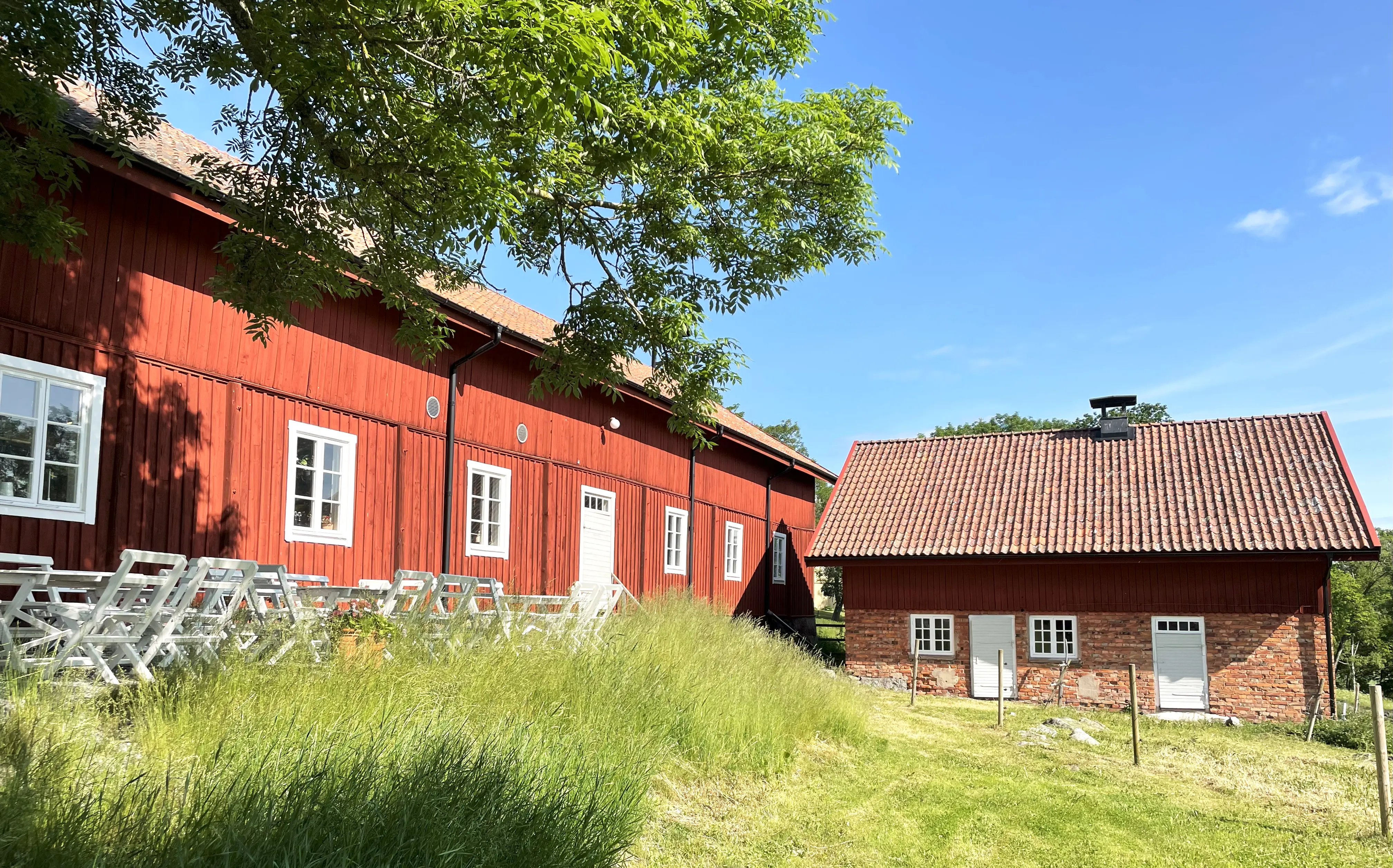 Landsberga. Äldre, rödmålat stall. Vita café-möbler står längs byggnaden. I vinkel mot den står ett gammalt hönshus i tegel.