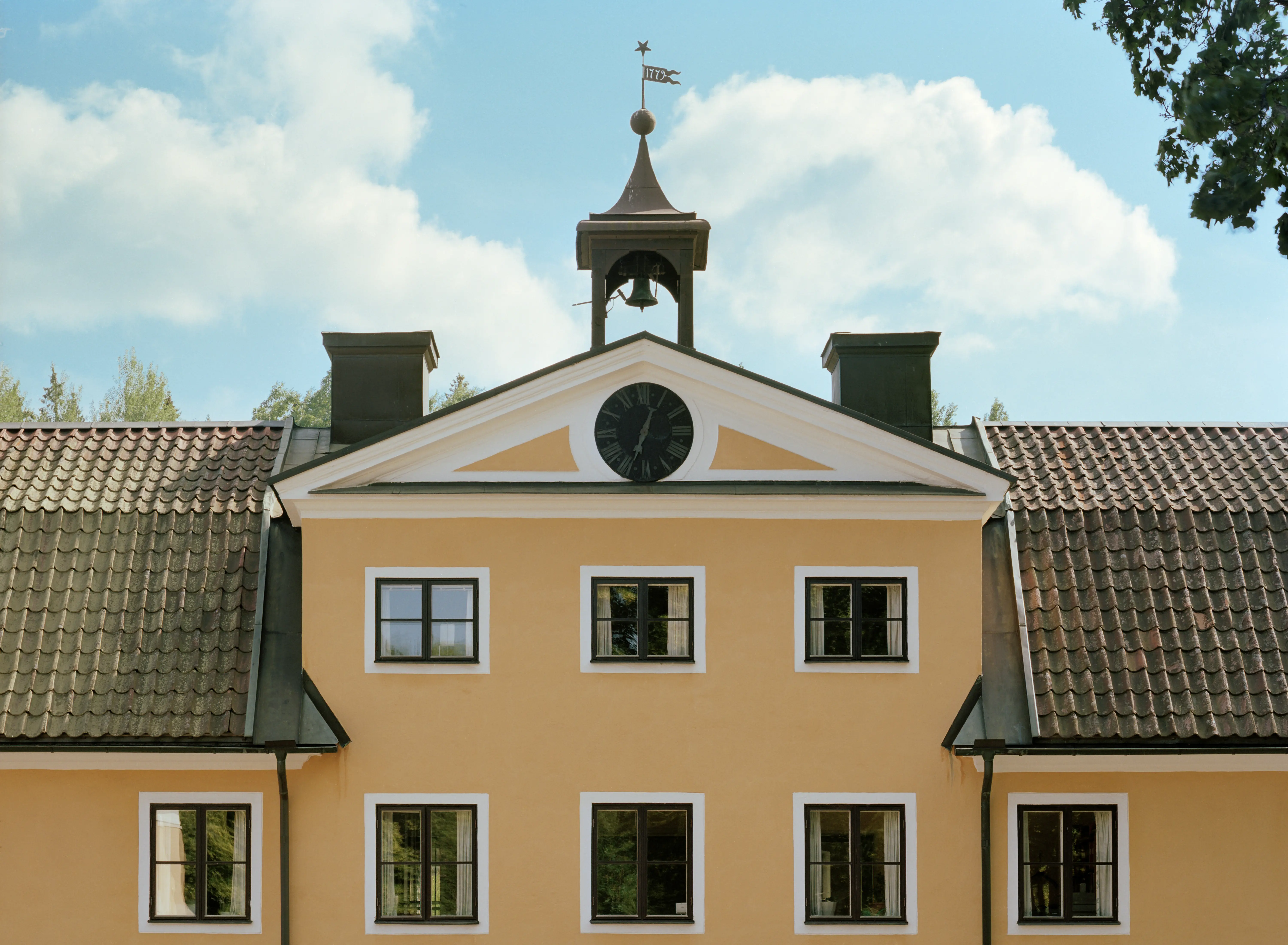 Klockhuset på Tumba bruk. Gulputsad byggnad med brutet tegeltak. På en mittgavel sitter en urtavla och på taket ovanför ett litet klocktorn.