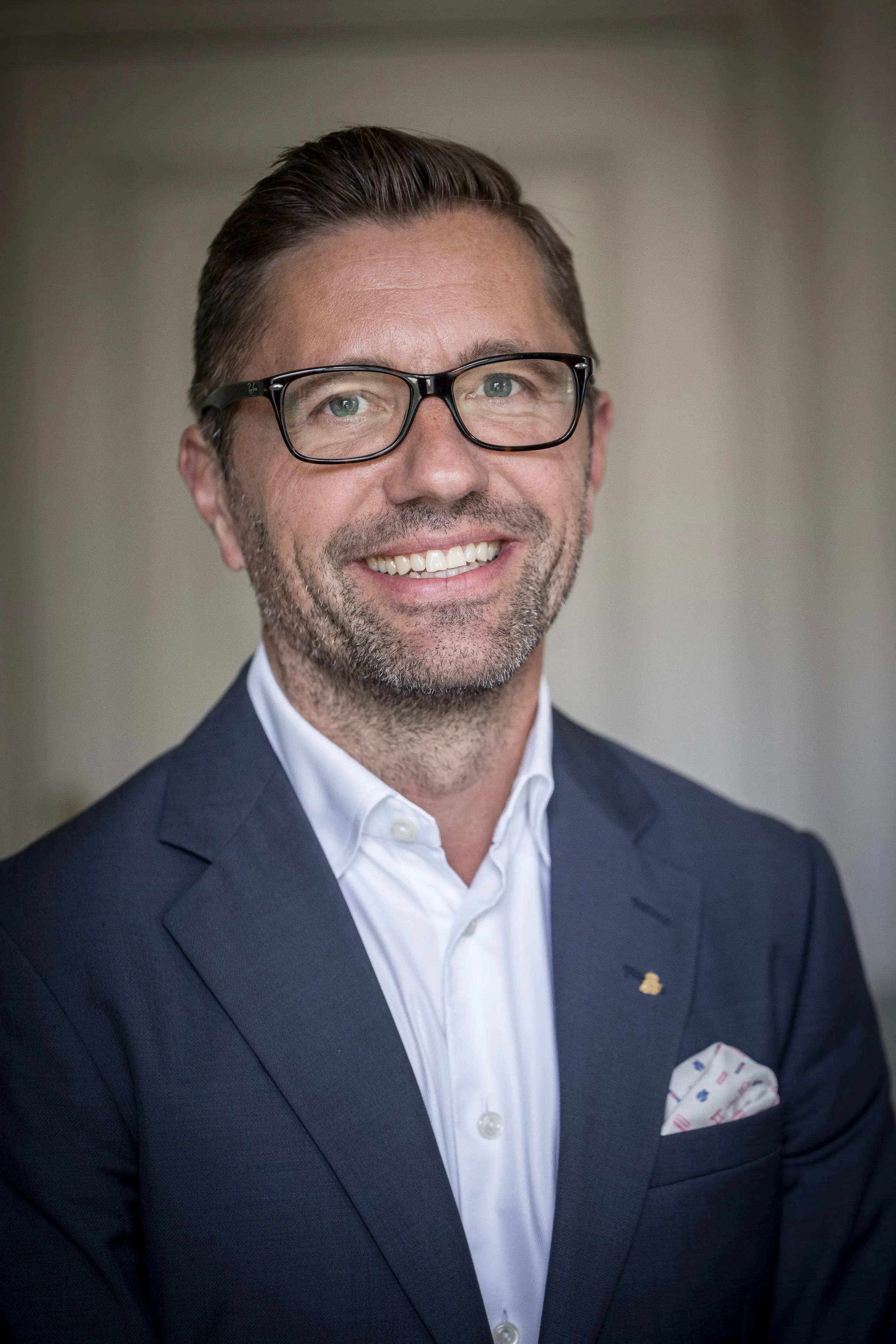Leende, glasögonprydd och skäggig man i marinblå kostym och vit skjorta.