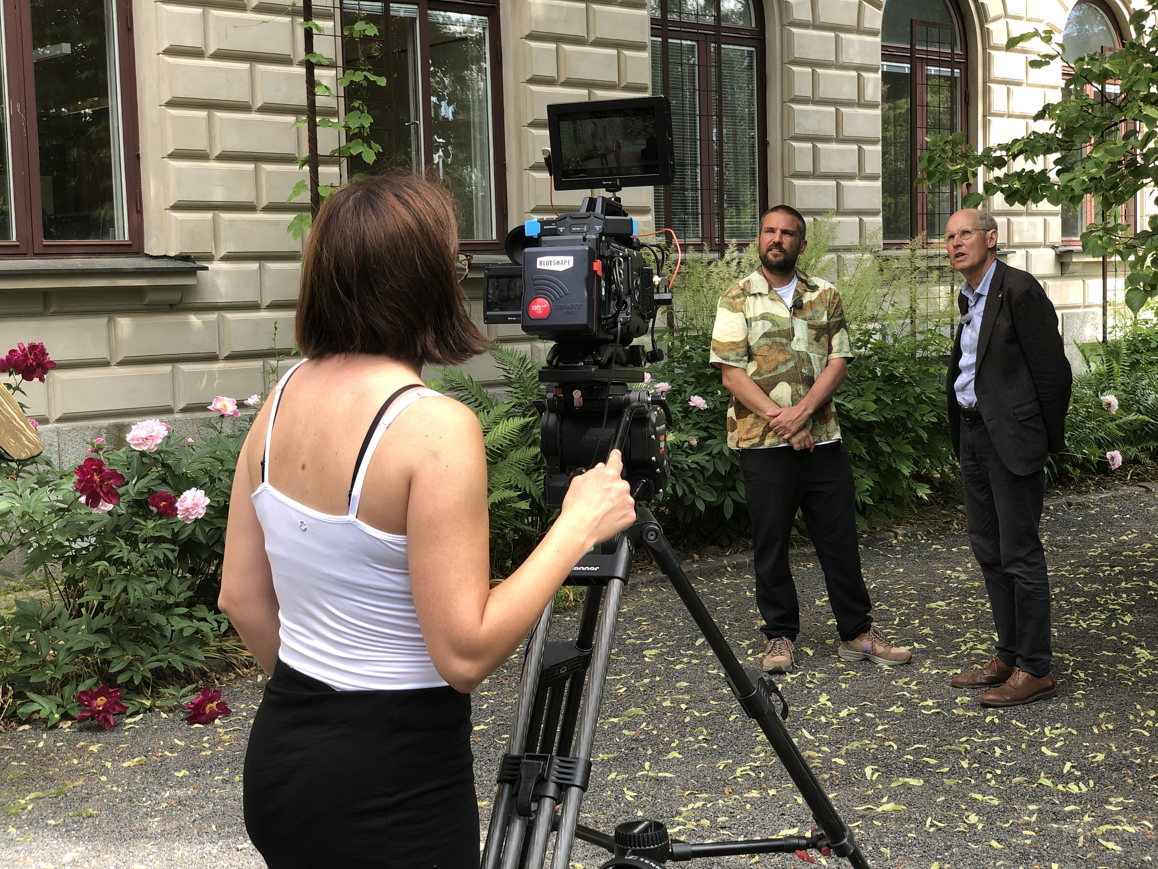 En person står framför en kamera och filmar två andra personer i en trädgård.